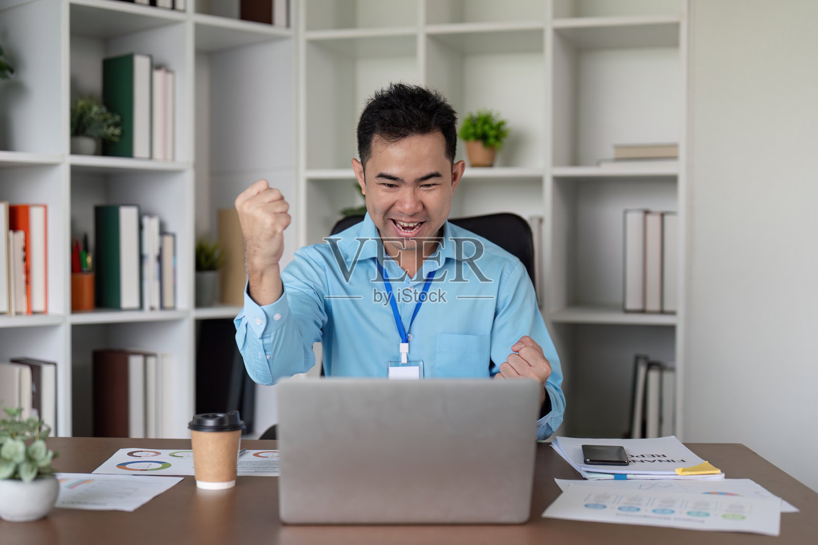 兴奋的亚洲商人在现代办公室的桌子上庆祝胜利,双拳举起,喜悦地使用笔记本电脑。照片摄影图片