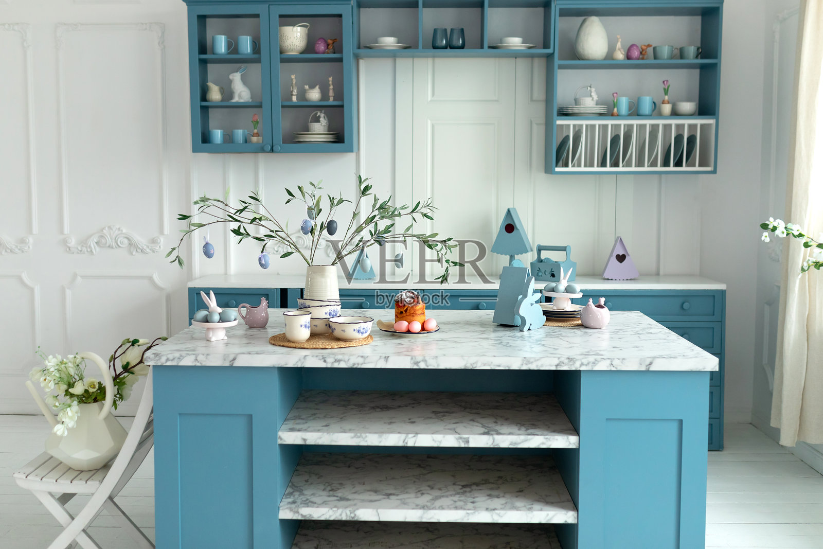 装有复活节彩蛋的橄榄树枝花瓶。蓝色厨房内饰,配有家具和复活节装饰。节日春季餐桌布置,复活节蛋糕和鸡蛋,厨房里的盘子。手工制作的潘纳托尼。照片摄影图片