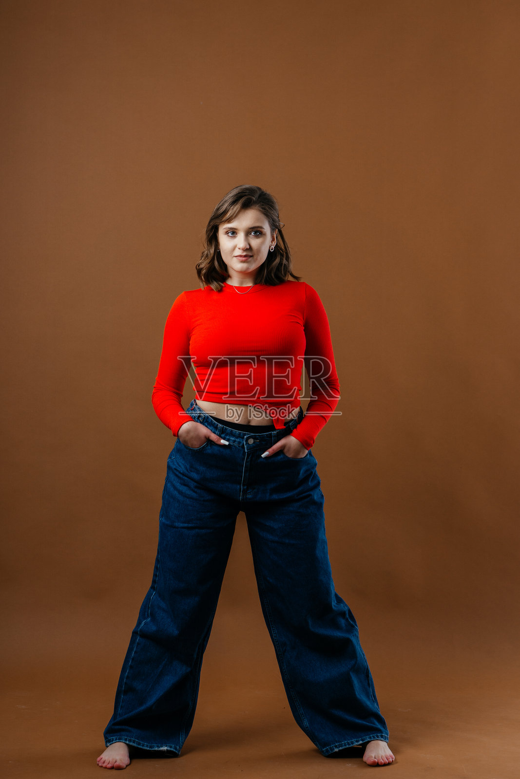 一位时尚的年轻女性穿着大胆的红色上衣和宽腿牛仔裤,在温暖的棕色背景前摆出动感而艺术的姿势。照片摄影图片