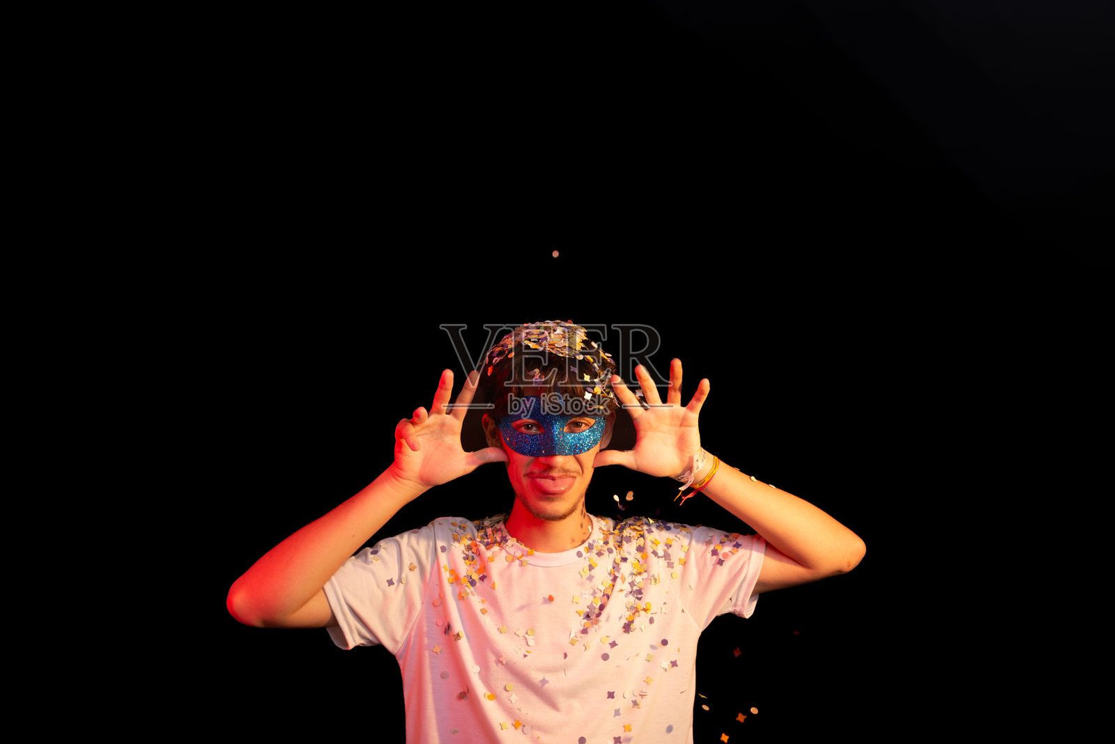 穿着白衬衫的年轻男子,戴着蓝色狂欢节面具,向镜头做手势,享受着五彩纸屑的乐趣。背景为黑色。照片摄影图片