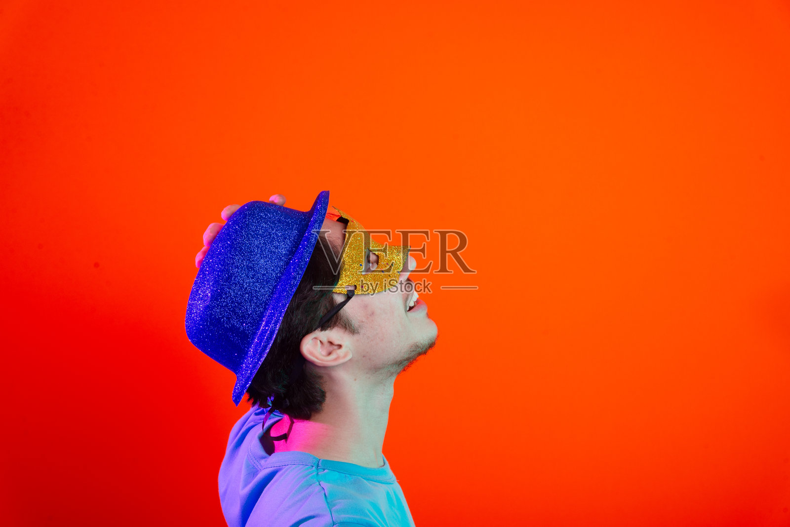 穿着蓝色衬衫、戴着狂欢节面具和蓝色帽子的年轻男子仰望。背景为霓虹橙色。照片摄影图片