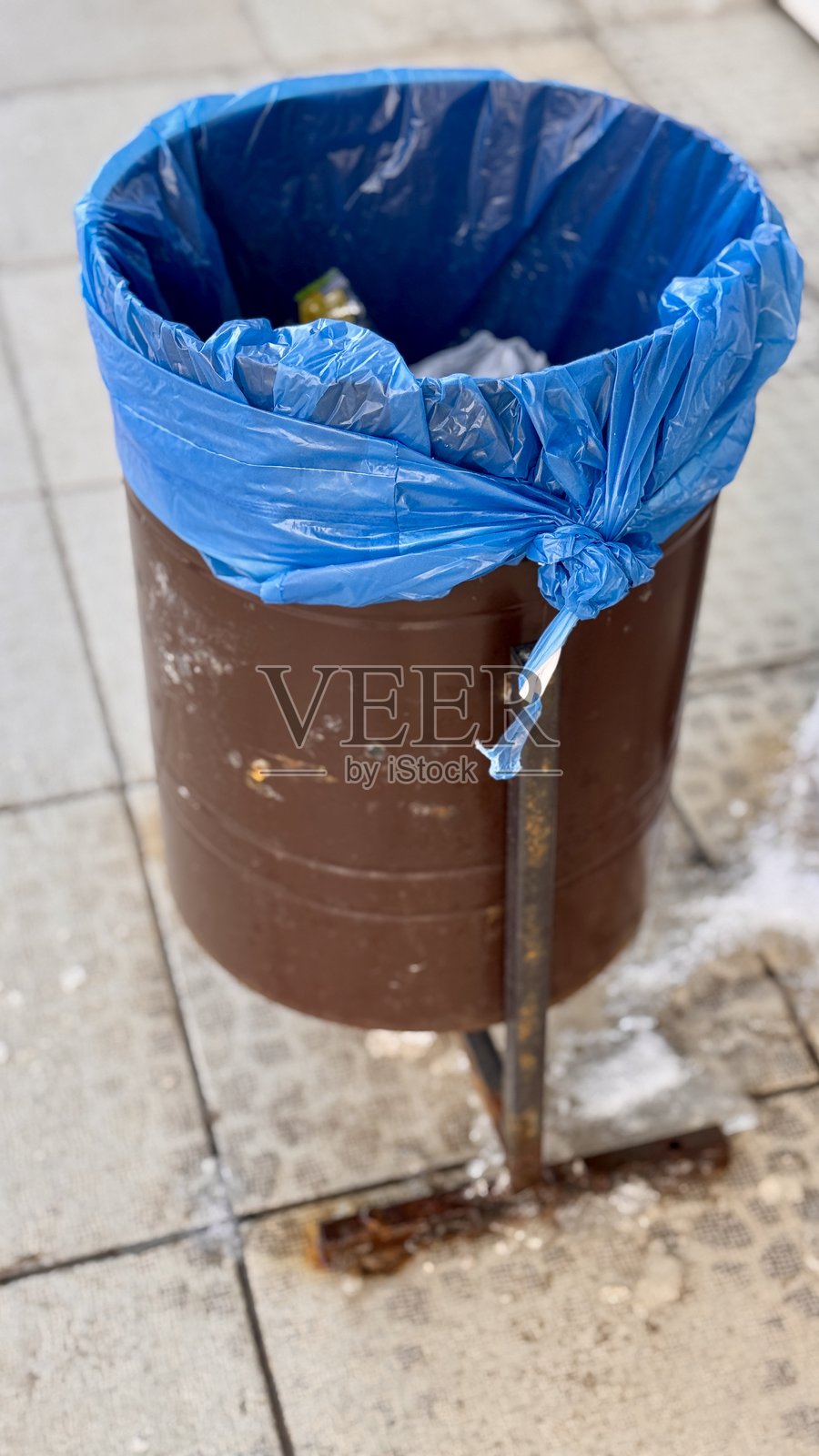 一个棕色的垃圾箱旁边放着一个蓝色的垃圾袋,安装在人行道上,靠近商店。特写。照片摄影图片