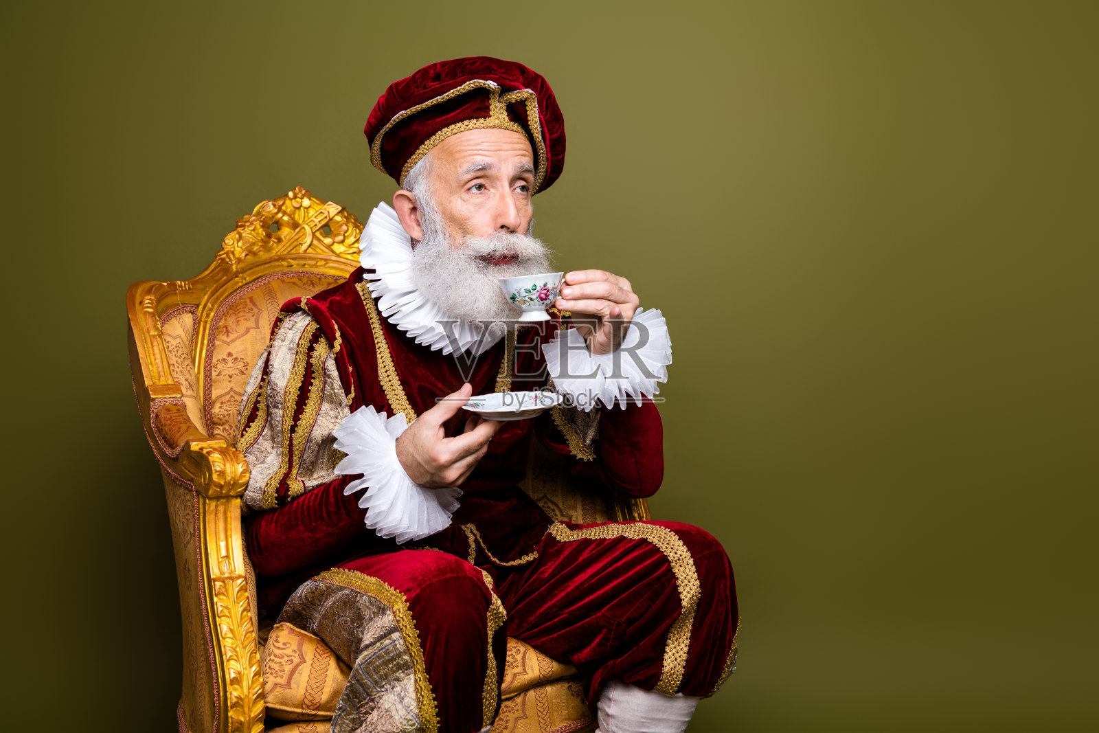 身穿历史服装的老年男子坐在华丽的椅子上品茶,背景为卡其色。照片摄影图片