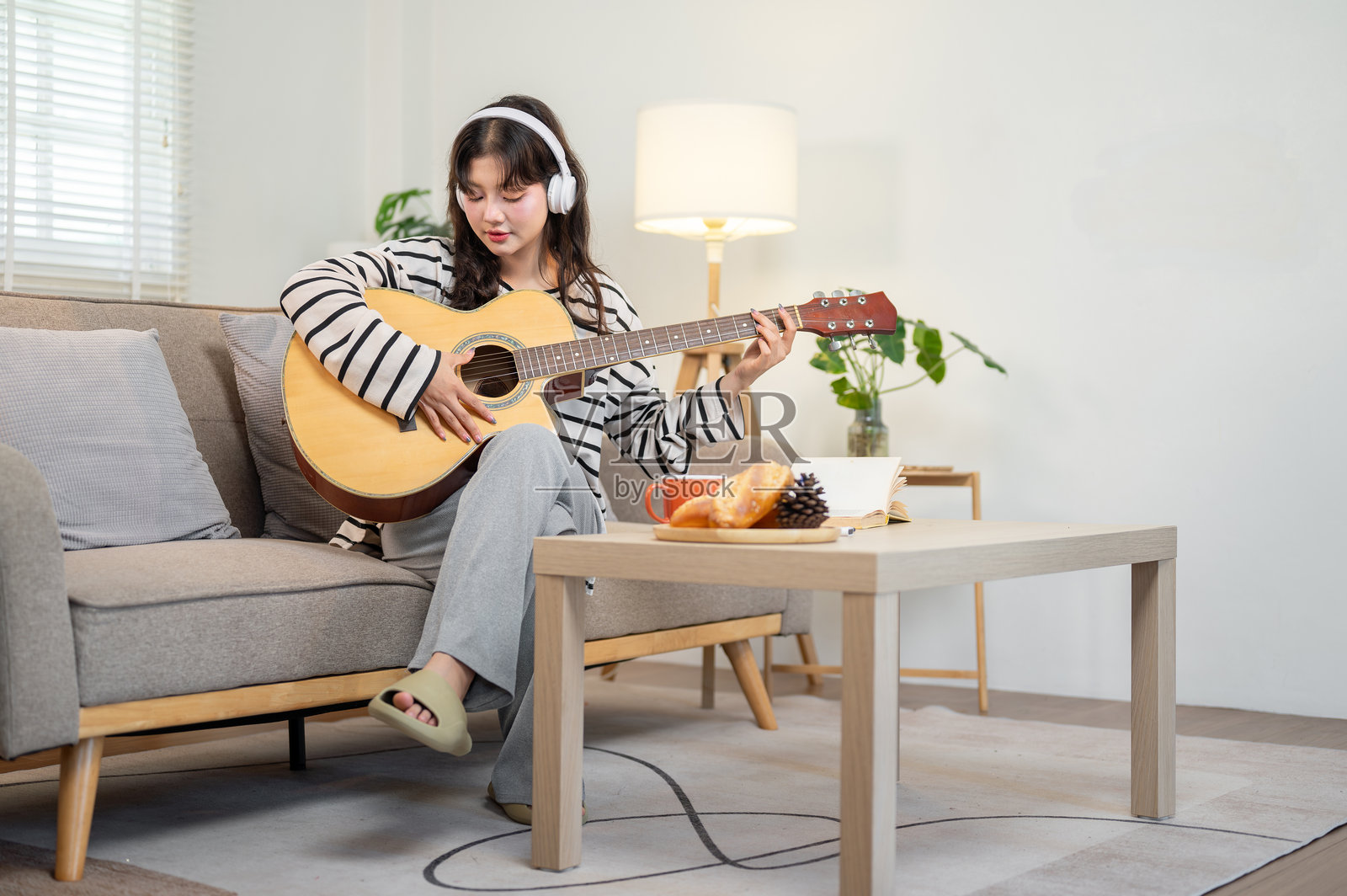 一位美丽、放松的亚洲女性坐在客厅的沙发上弹吉他。照片摄影图片