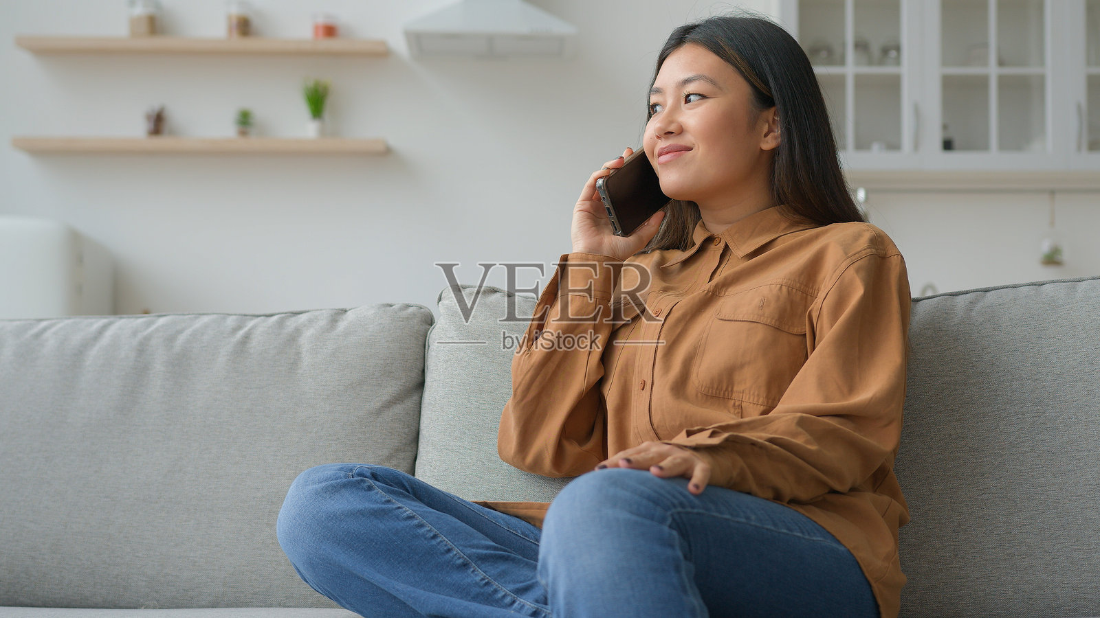 无忧无虑的亚洲女性在厨房的沙发上打手机,微笑着进行远程通话。照片摄影图片