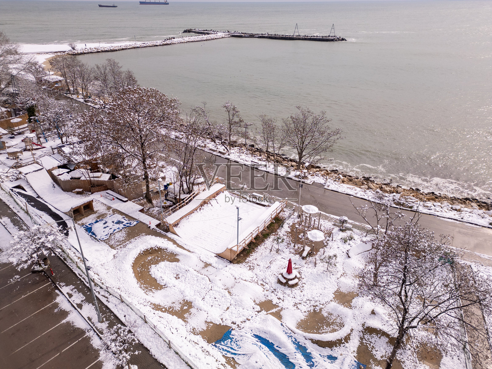 冬季海滨景观的空中视角,覆盖着雪。宁静的海岸场景,有一条道路、树木和靠近平静海洋的小房子。阳光在水面上反射,增添了温暖。照片摄影图片