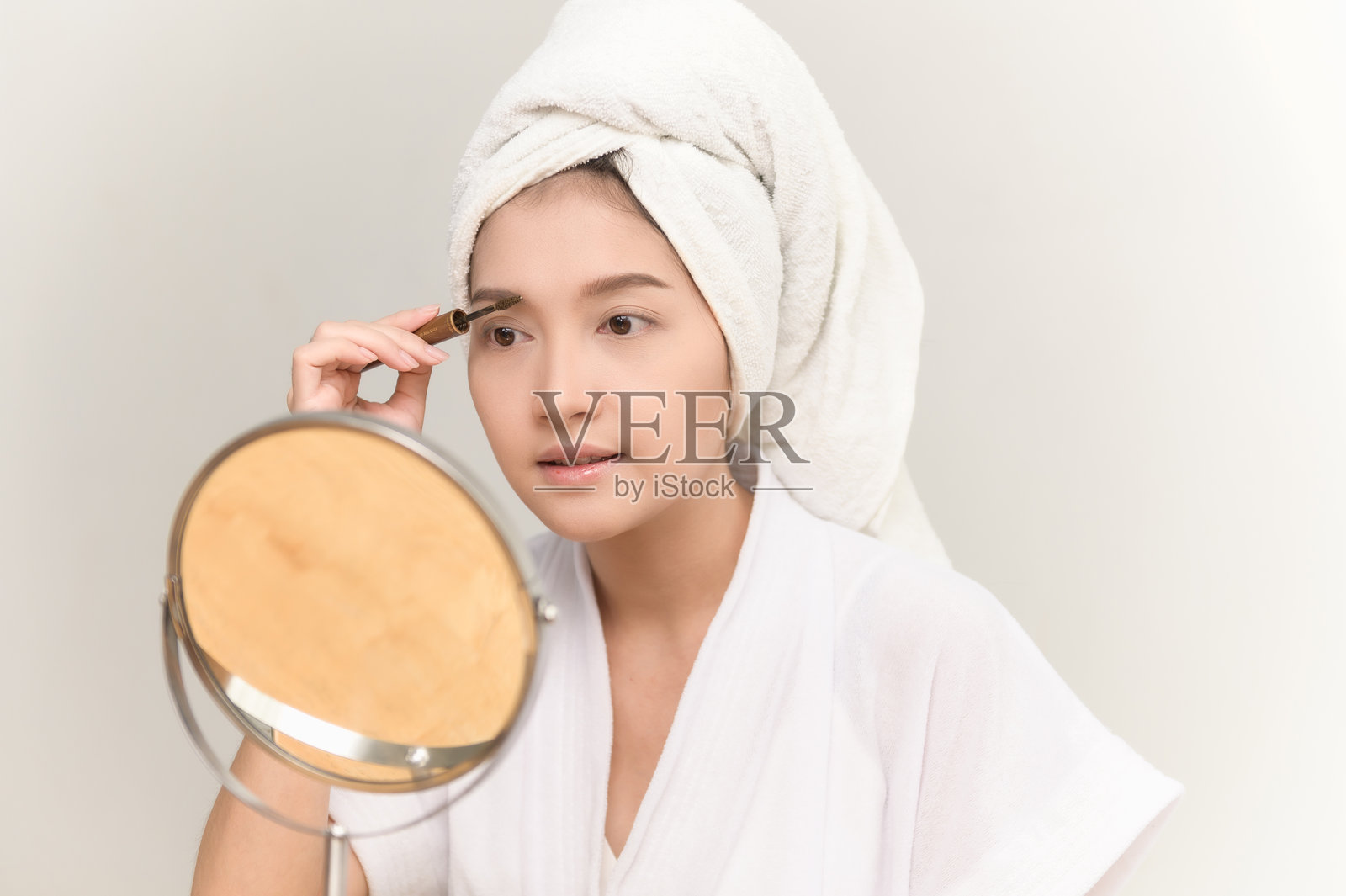 一位年轻的亚洲女性头上裹着毛巾,正对着镜子给自己的眉毛化妆。美容程序,护肤,自我护理,化妆教程,自然妆容,美容护理概念。照片摄影图片