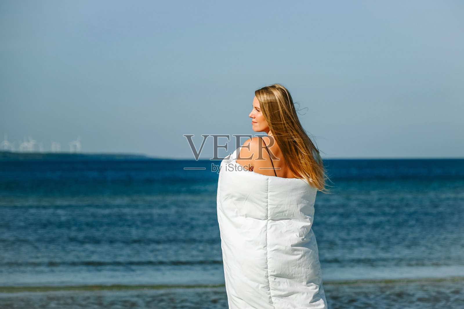 身穿白色毛毯的女人在沙滩上沿海边散步。照片摄影图片