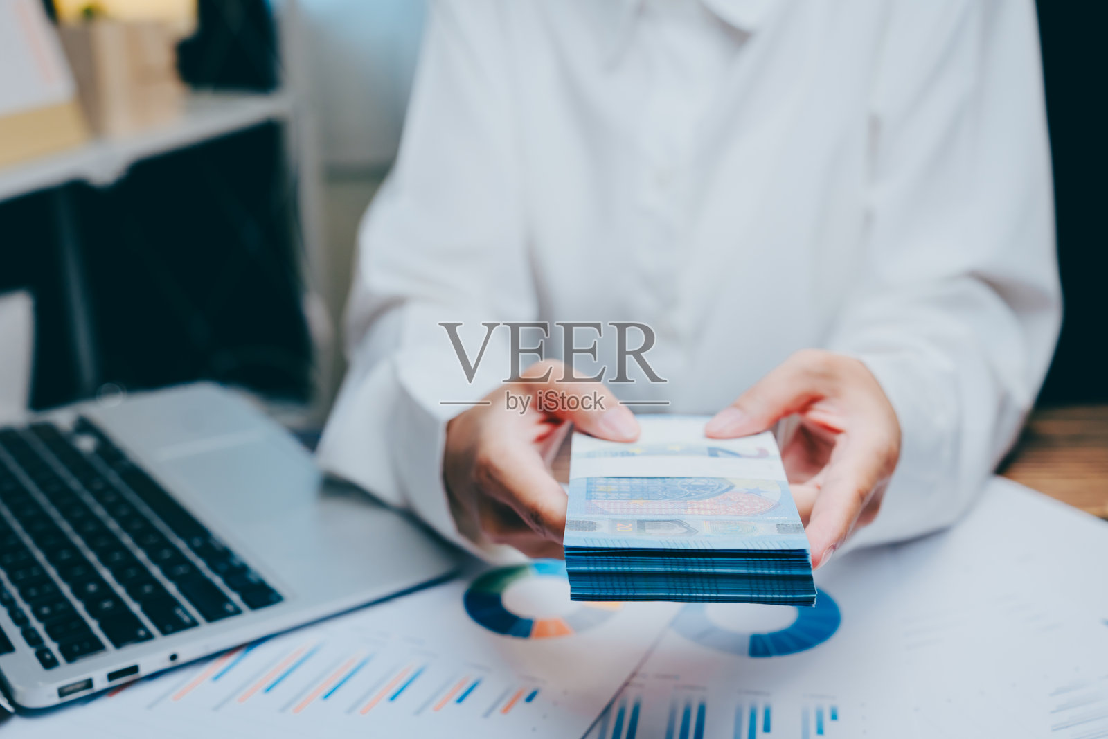 手握现金堆叠,旁边有笔记本电脑和财务图表,商业概念照片摄影图片