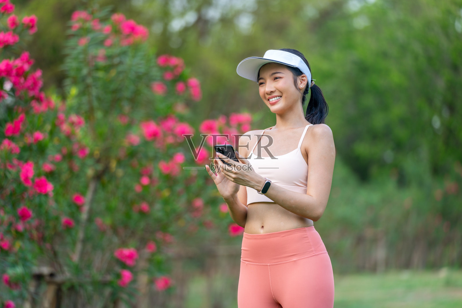 年轻亚洲女性穿着运动服,戴着遮阳帽,在盛开的公园里锻炼时使用手机照片摄影图片