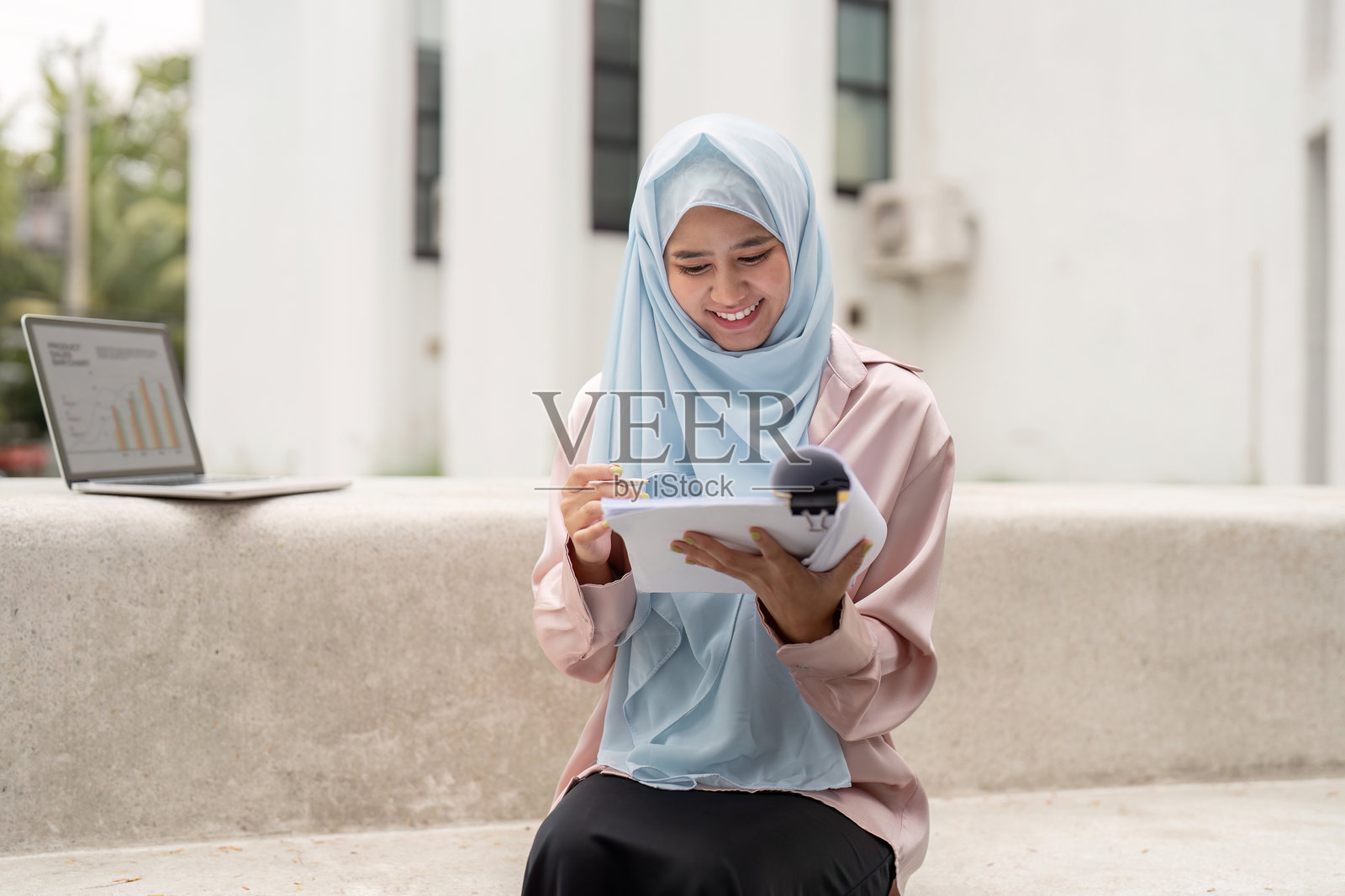 快乐的穆斯林女商人在户外城市环境中坐在笔记本电脑旁,分析平板上的数据。强调现代创业和文化多样性。照片摄影图片