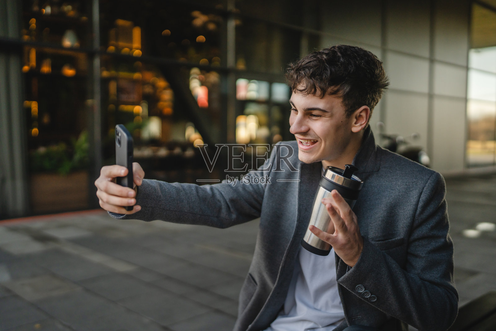 年轻男子坐在长椅上喝咖啡,正在用手机进行视频通话照片摄影图片