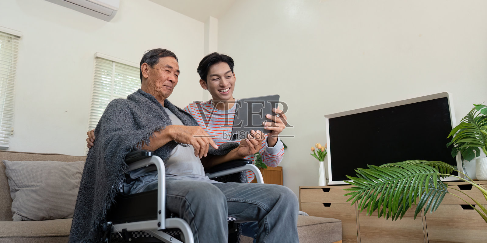 儿子在家中与坐在轮椅上的老父亲共享快乐时光,一起使用平板电脑。照片摄影图片