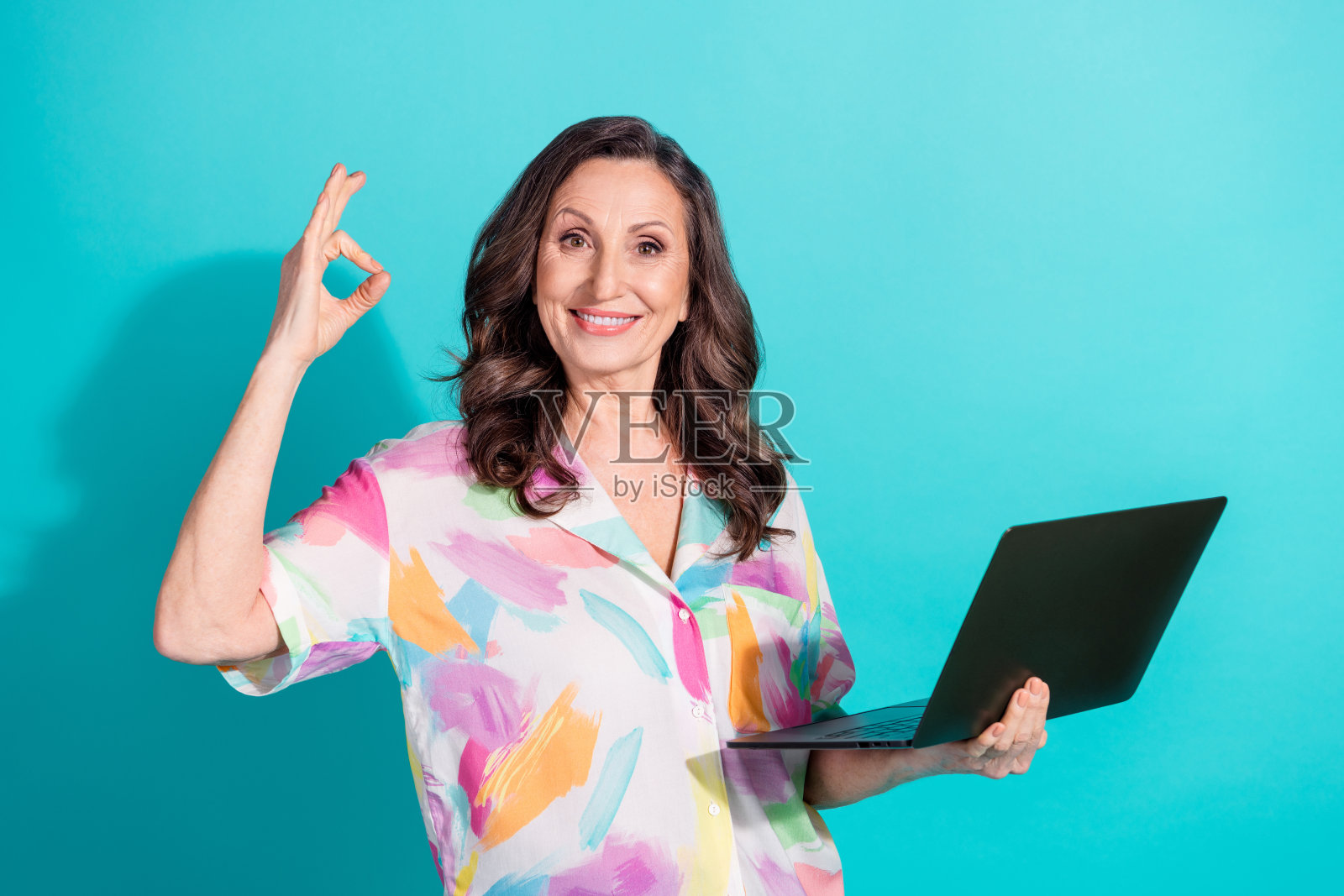 漂亮的退休女性手持笔记本电脑,做出“好”的手势,穿着时尚多彩的服装,背景为青色。照片摄影图片
