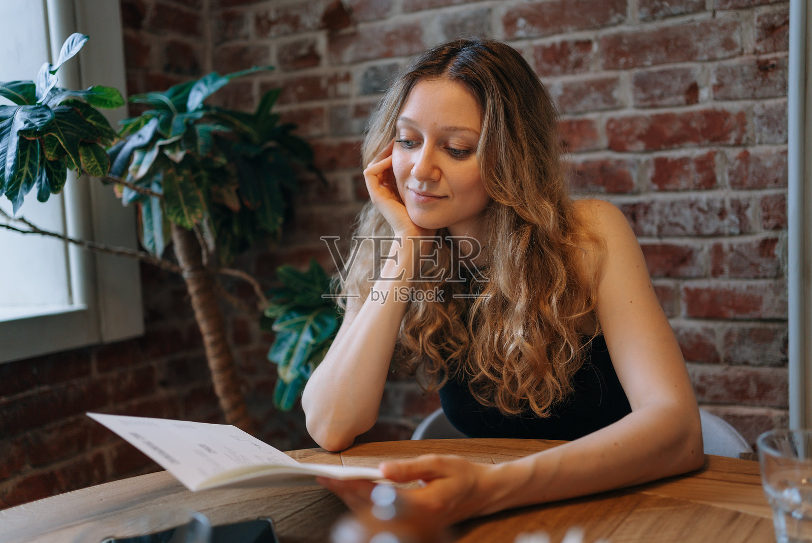 女人在舒适的咖啡馆里阅读菜单,坐在木桌旁,背景是砖墙。照片摄影图片