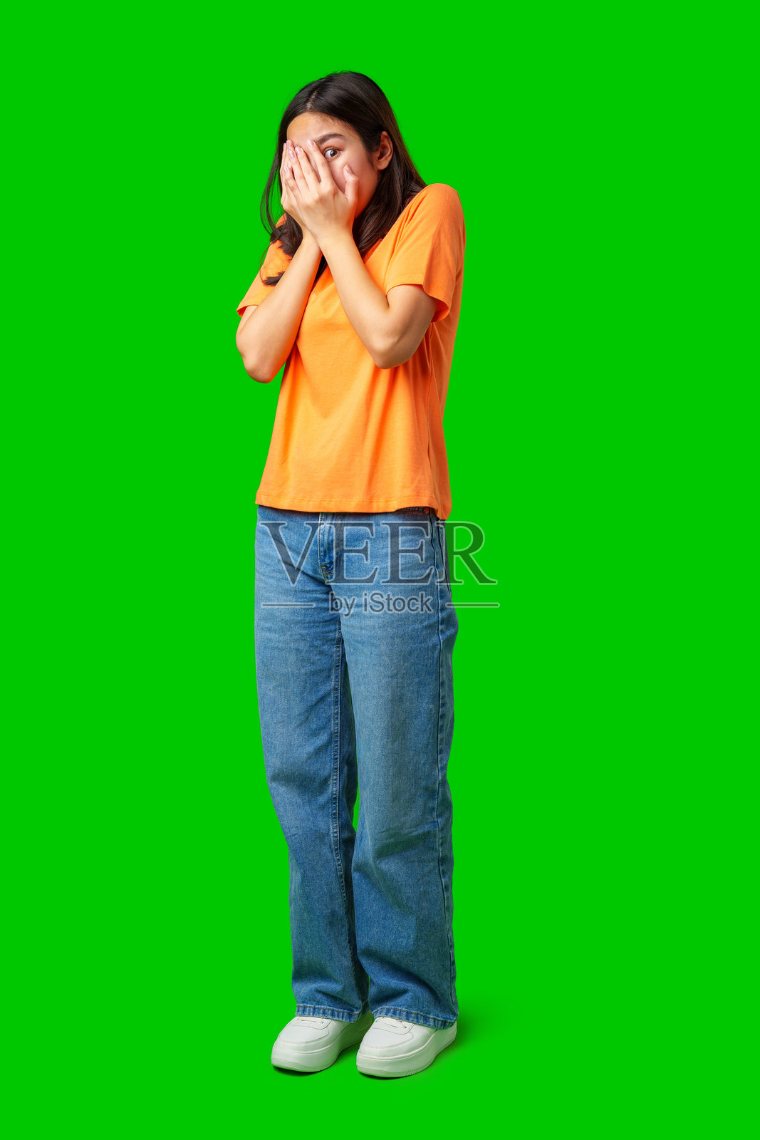 年轻女性穿着橙色衬衫,长发披肩,用双手遮住脸,站在绿色背景前。照片摄影图片