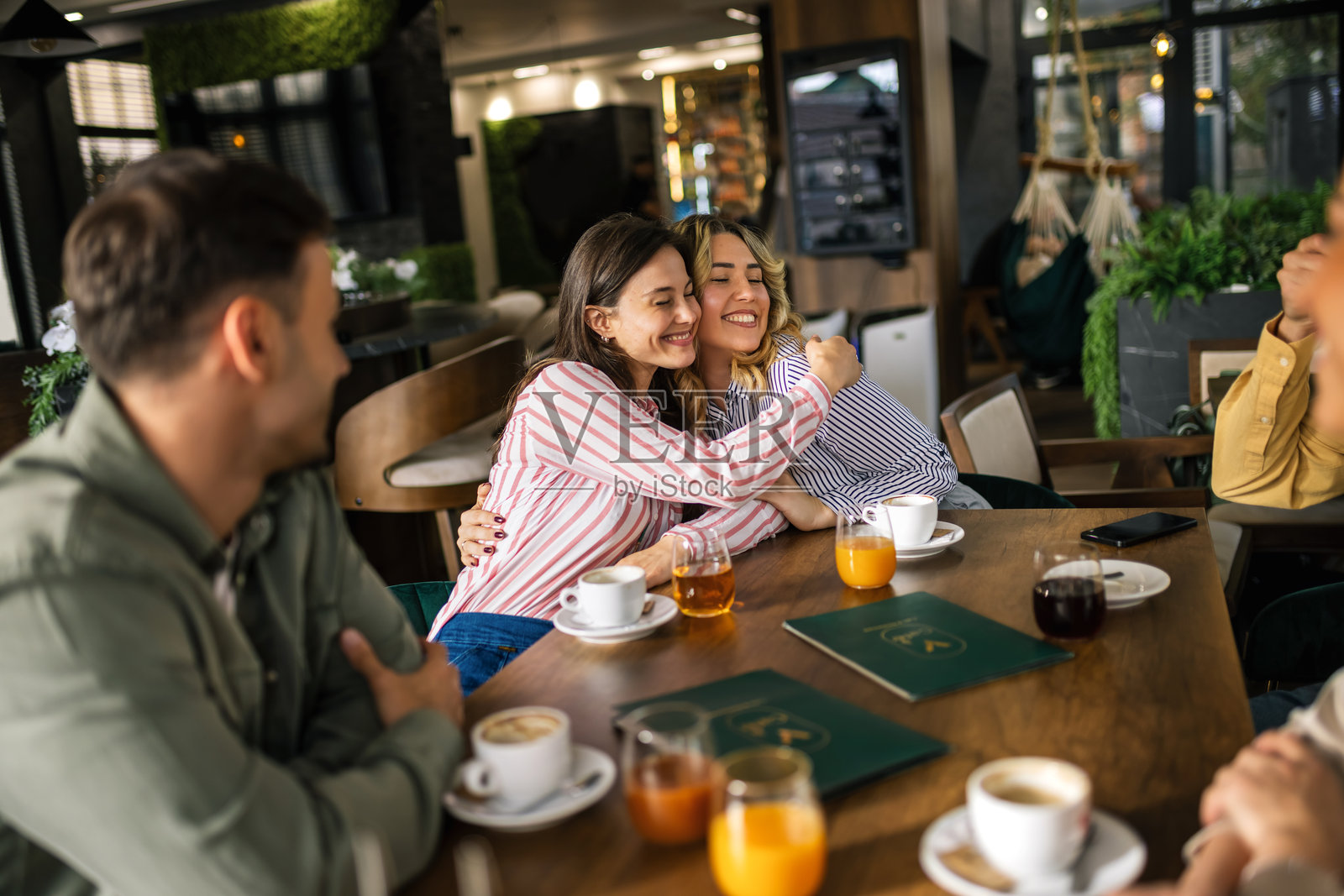 一群年轻人坐在现代咖啡馆的桌子旁,享受着彼此的时光,手中拿着咖啡杯和新鲜榨汁的杯子。照片摄影图片