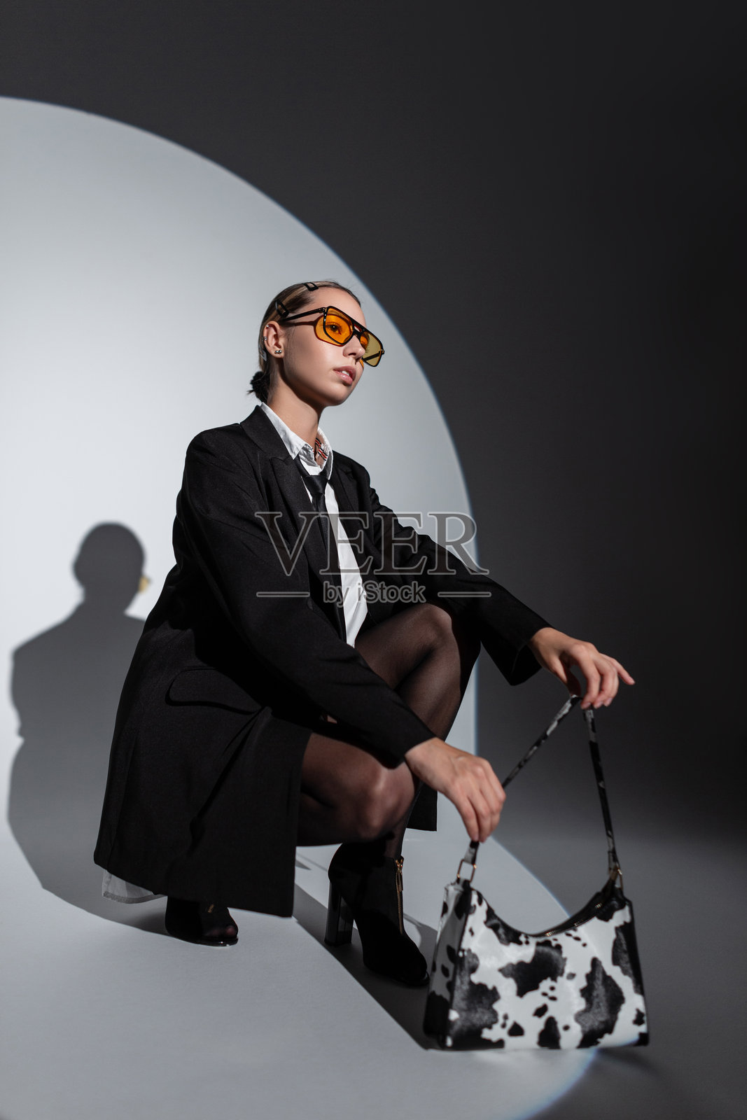 时尚迷人的女经理穿着时尚的衣服,戴着黄色眼镜,身穿西装外套、衬衫和领带,手提手袋,坐在工作室里摆姿势。照片摄影图片