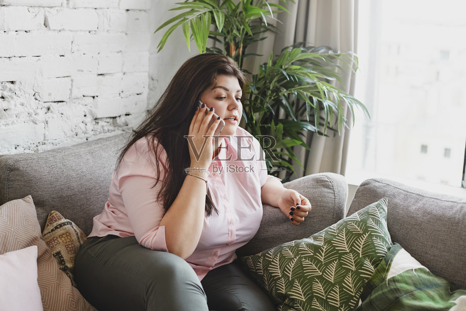 坐在窗边沙发上的迷人年轻棕发丰满女性打电话,正在与外卖服务沟通,点购面条。肥胖、人与科技的概念。照片摄影图片