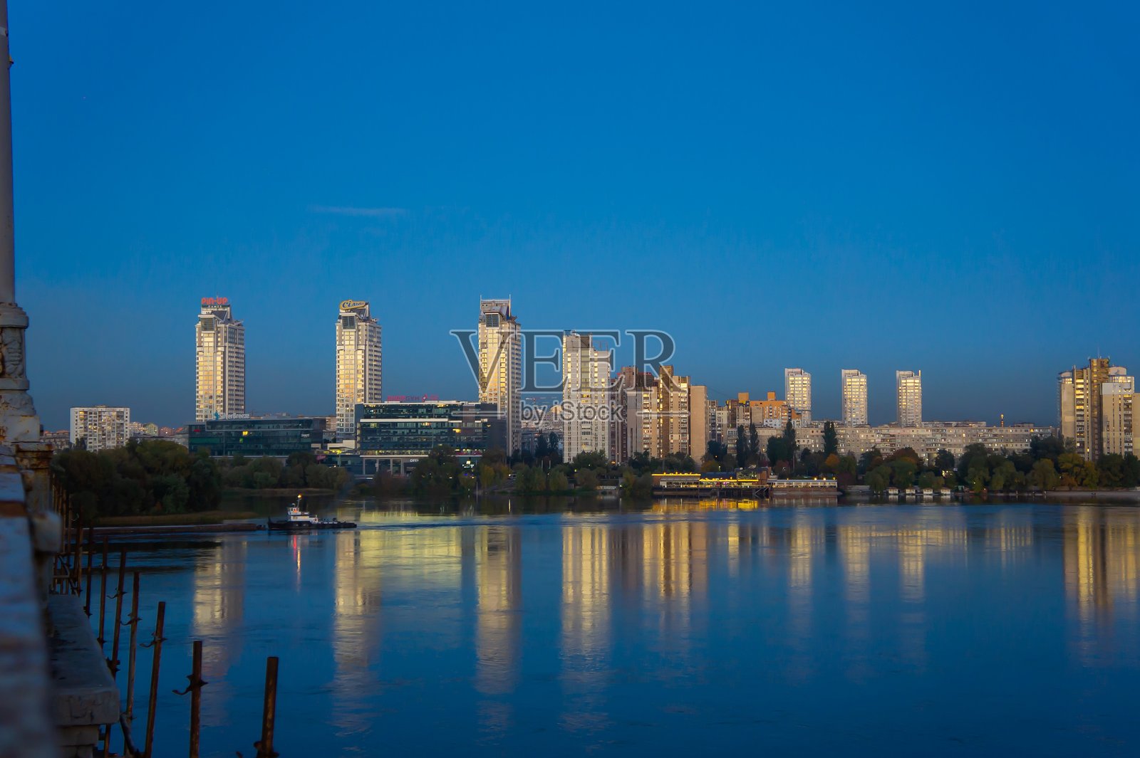 黄昏时分的现代城市景观,灯光闪烁的建筑倒映在宁静的水面上,前景的树木剪影与亮起的建筑形成对比。照片摄影图片