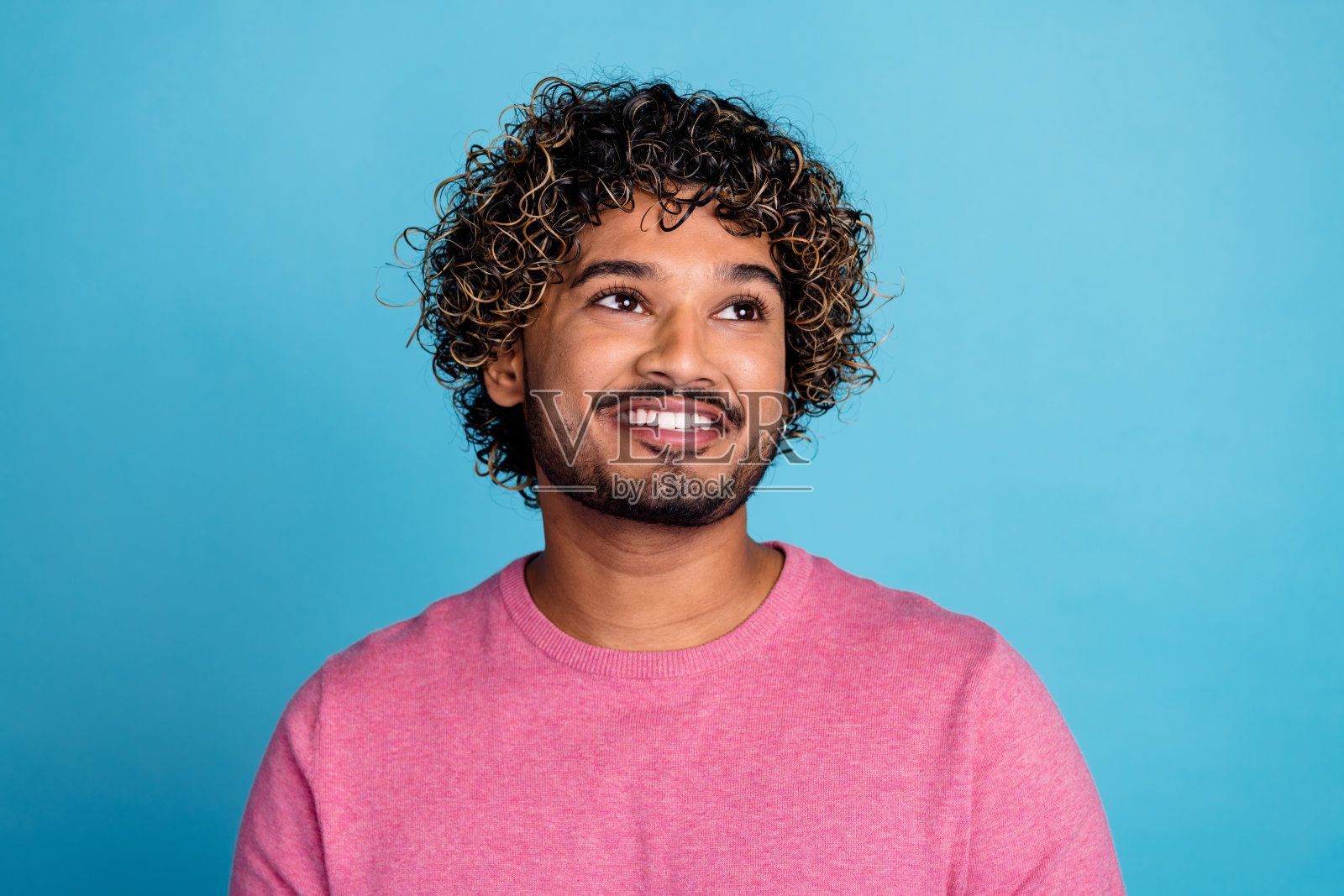 一位穿着粉色毛衣、卷发的年轻男子在明亮的蓝色背景下微笑着仰望。照片摄影图片