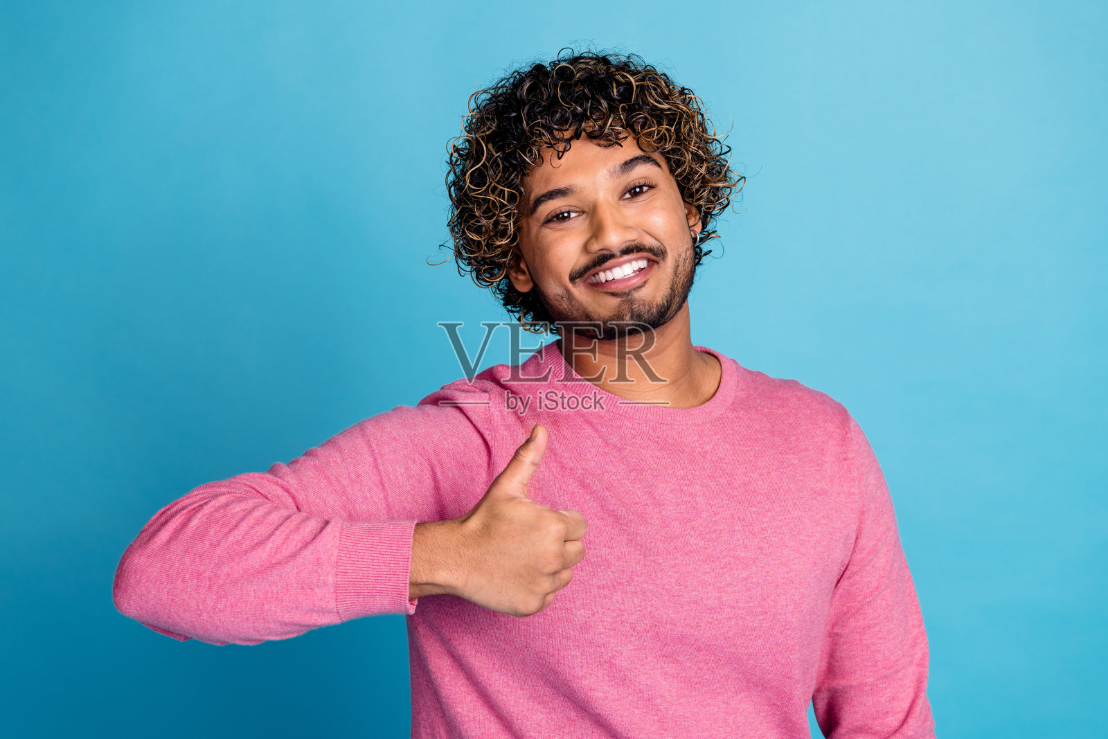 年轻的微笑男士穿着粉色休闲服,面带愉快的表情,竖起大拇指,背景为鲜艳的蓝色。照片摄影图片