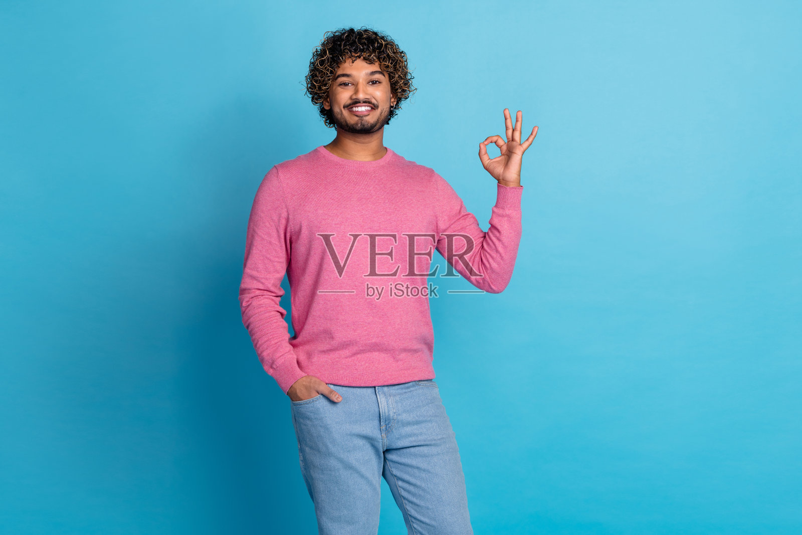 穿着粉色毛衣的时尚年轻男子在蓝色背景前做出“好的”手势照片摄影图片