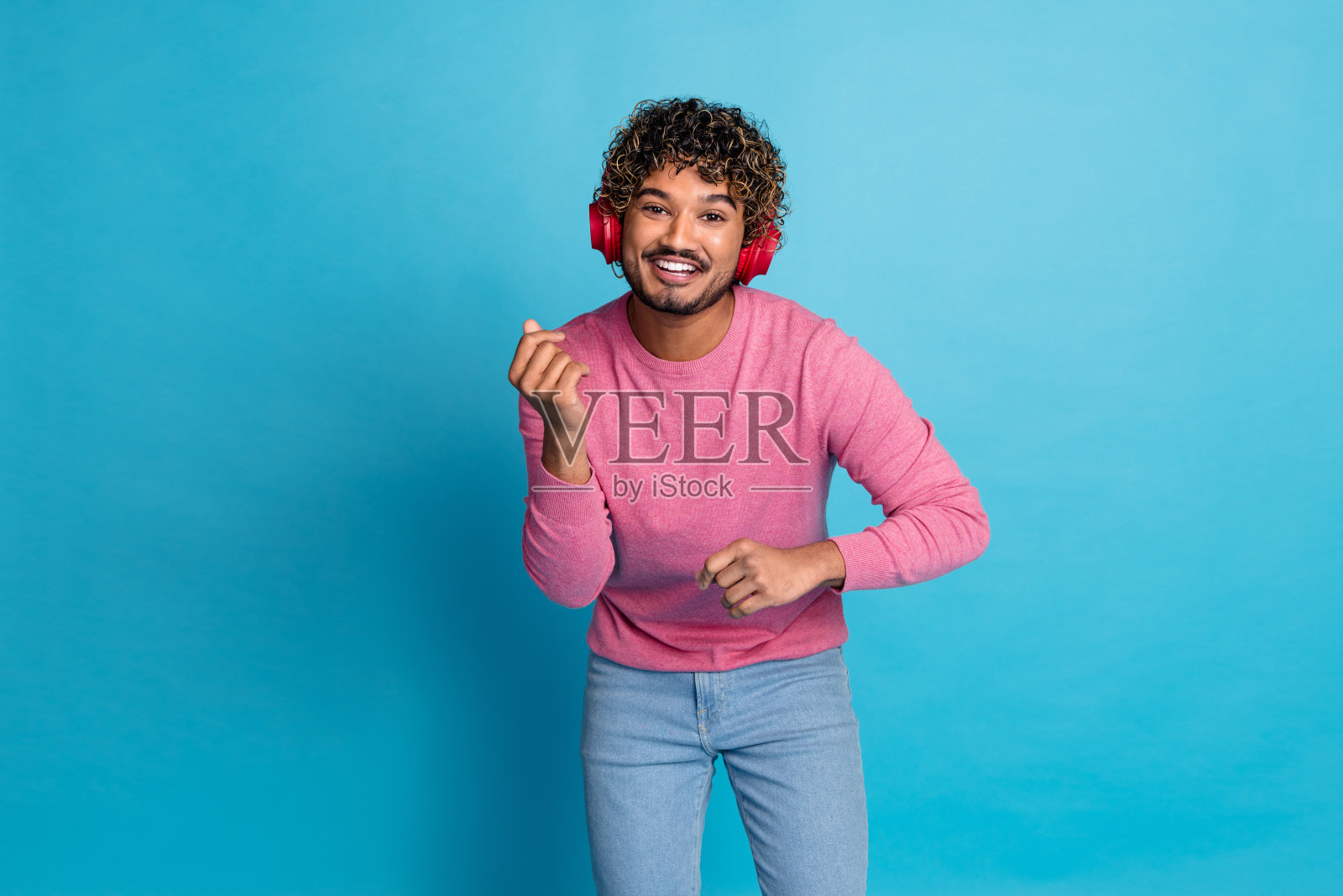 年轻男子穿着休闲粉色服装,戴着耳机,在鲜艳的蓝色背景下享受音乐照片摄影图片