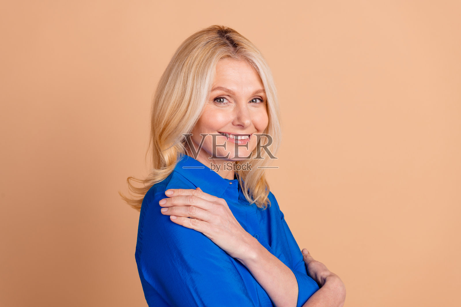 一位微笑的成熟的女性,穿着蓝色衬衫,金色头发,背景为米色,展现出自信。照片摄影图片