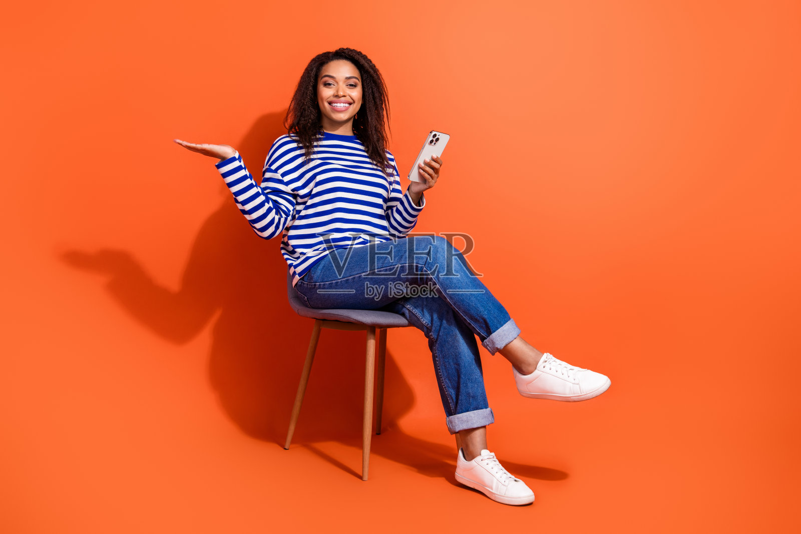 可爱的女孩穿着条纹衬衫,手持现代设备发短信,展示空白的手臂,背景为橙色。照片摄影图片