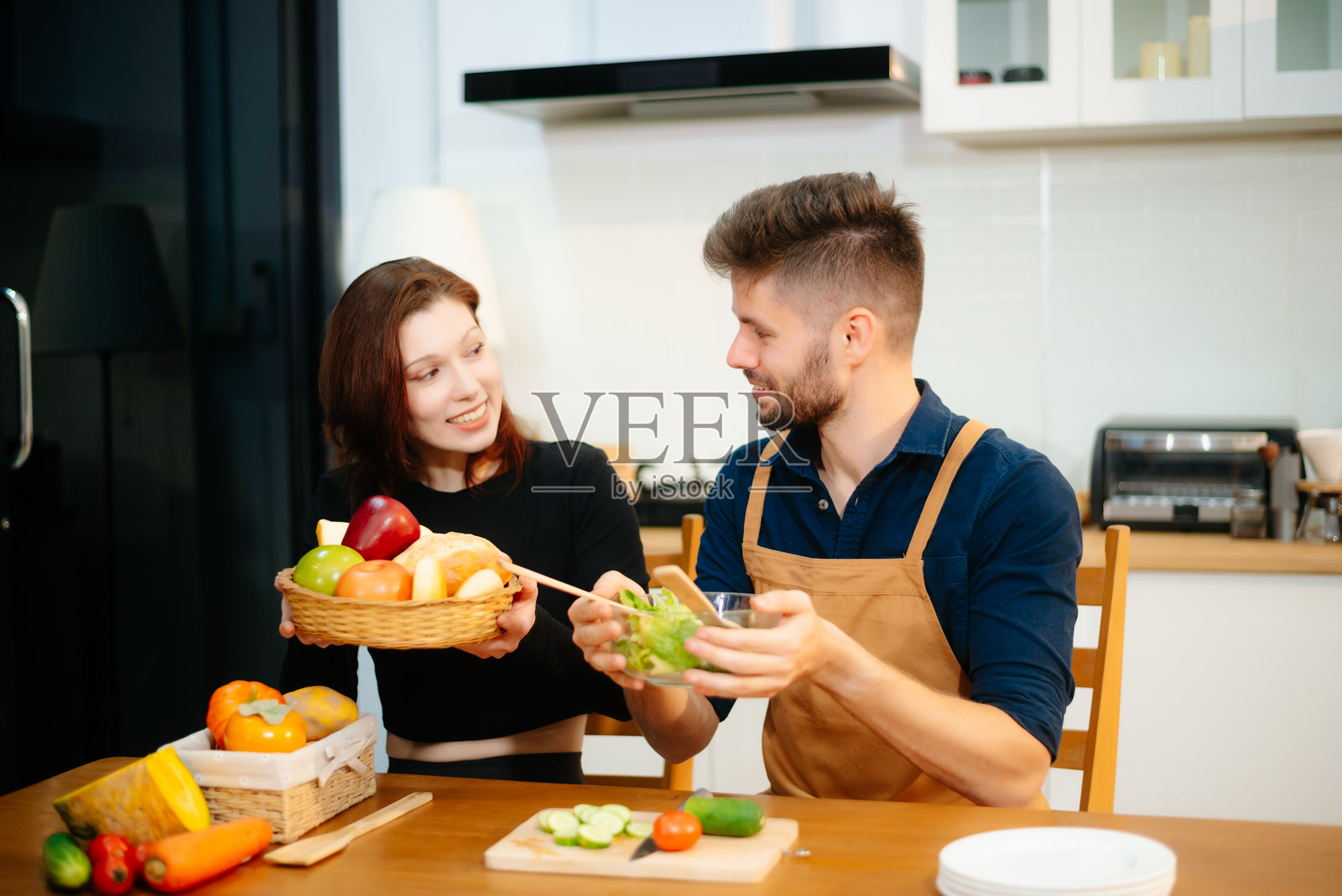 年轻情侣在温馨的厨房里一起享受烹饪新鲜的蔬菜、水果和面包。照片摄影图片
