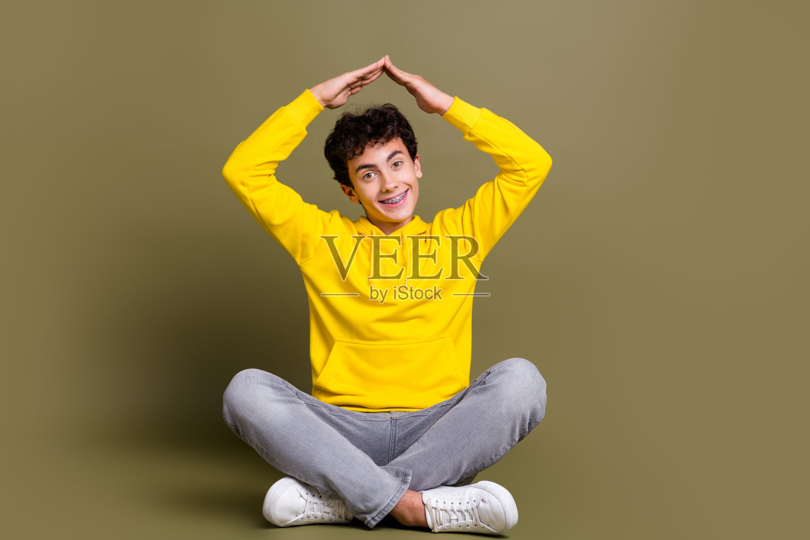 微笑的年轻男子穿着黄色连帽衫,盘腿坐在卡其色背景上,开心地摆姿势。照片摄影图片