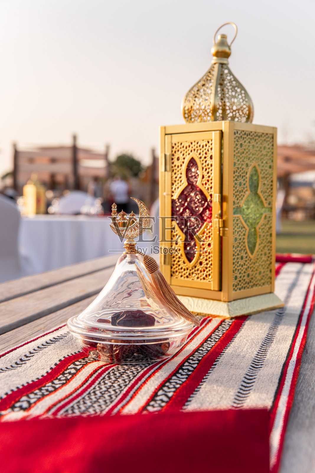 优雅的玻璃碗里装着枣子,旁边是金色的阿拉伯灯笼,摆放在阿布扎比户外花园草坪上的节日开斋餐桌上,夕阳下的庆祝场景。照片摄影图片