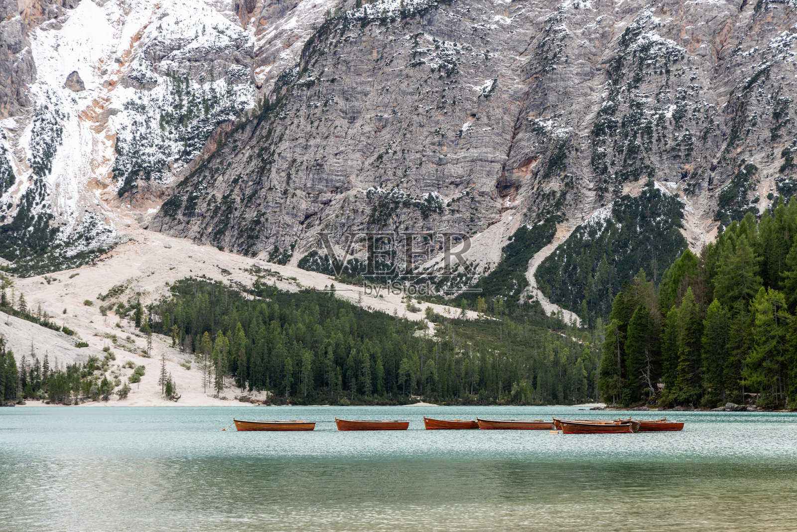 木船静静地停靠在布拉耶斯湖碧绿的水面上,四周被覆盖着雪的陡峭悬崖和茂密的阿尔卑斯森林环绕,位于意大利北部。照片摄影图片