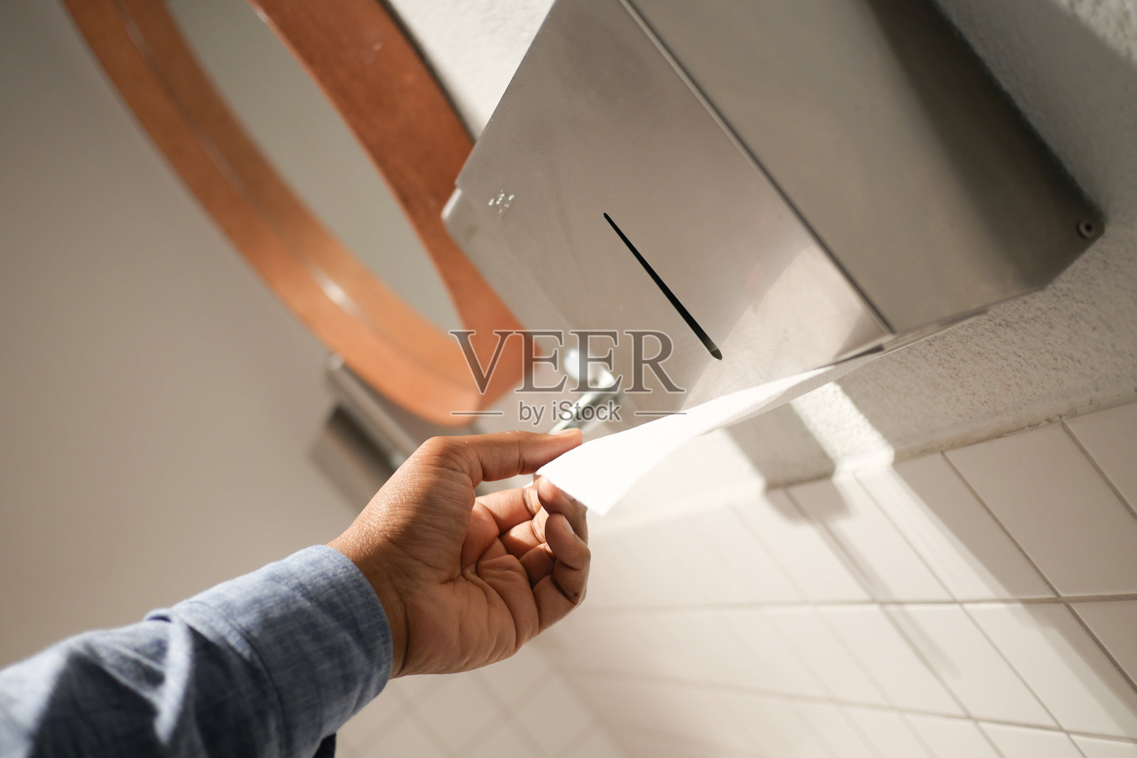 在卫生洁净的公共洗手间中,手正在使用纸巾分配器,周围是干净的白色瓷砖。照片摄影图片