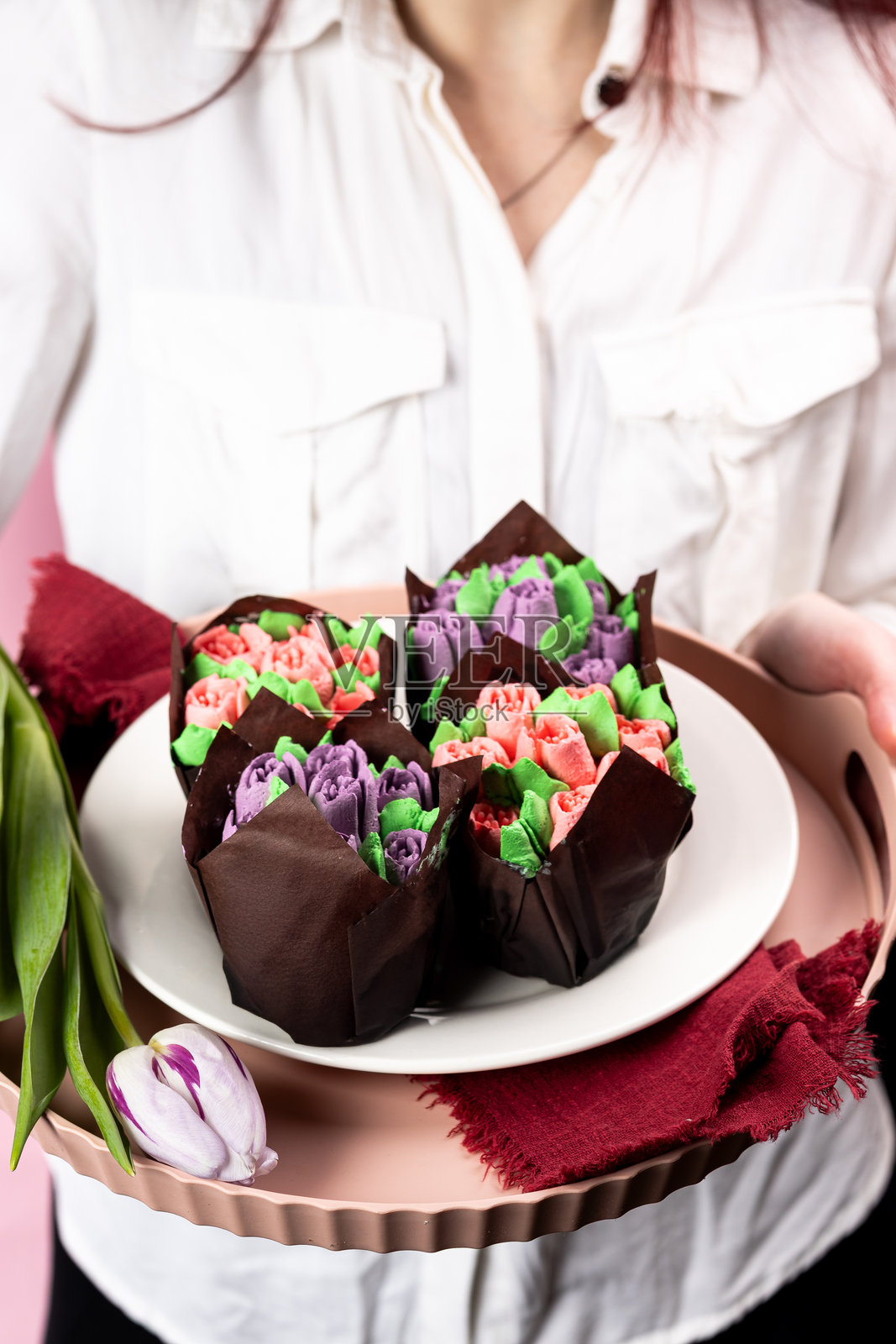 穿着白衬衫的女人手持粉色托盘,上面放着形状如粉色和紫色郁金香的奶油松饼,母亲节杯子蛋糕,三月八日的概念,奶油松饼,奶油郁金香,母亲节祝福照片摄影图片