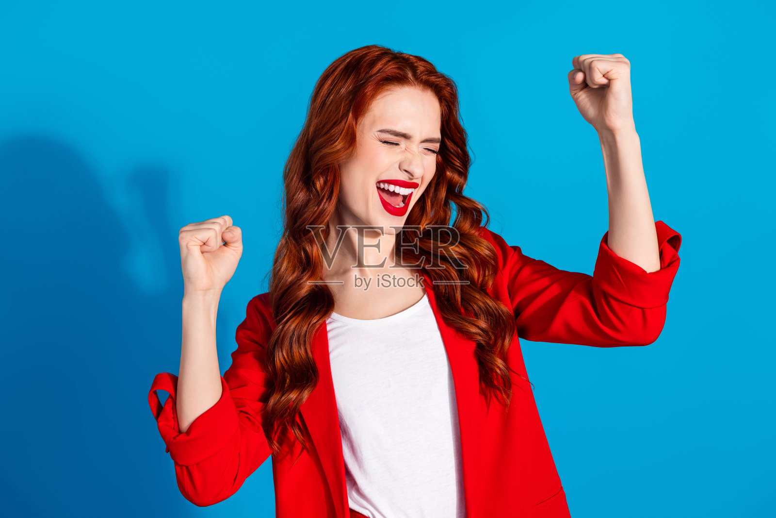快乐、开朗、迷人的女孩穿着红色时尚服装,双手握拳,背景为蓝色。照片摄影图片