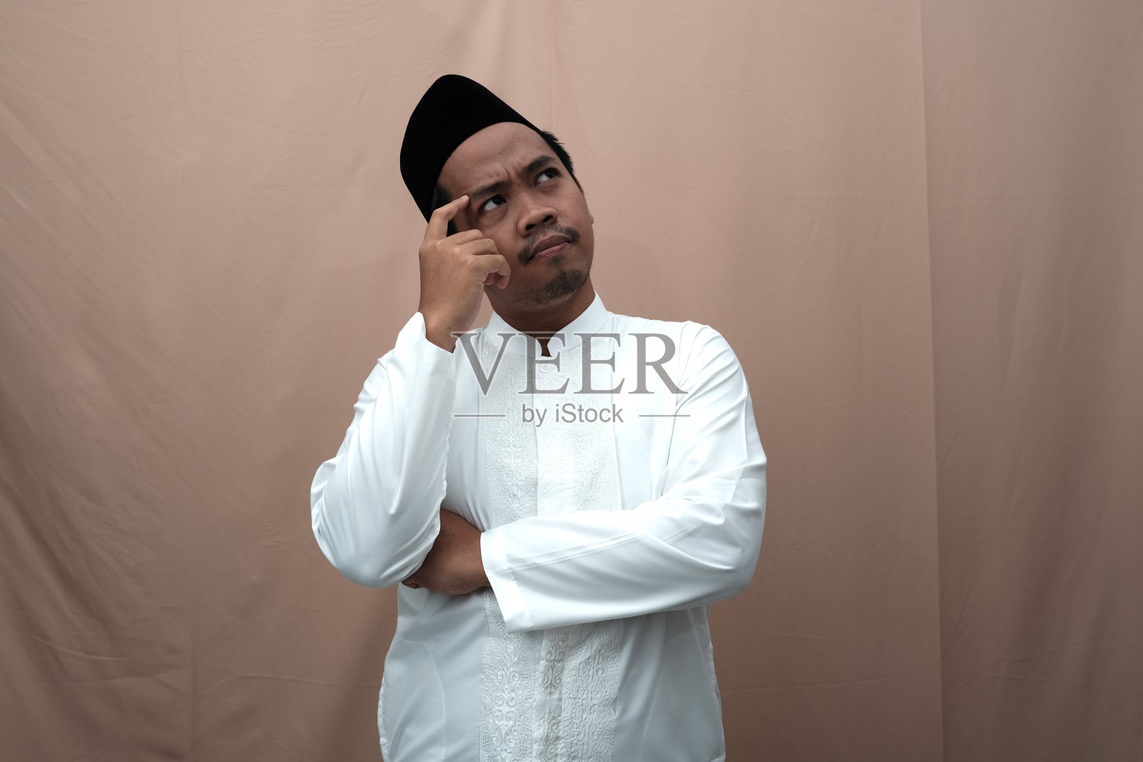 沉思的亚洲穆斯林男子,身穿传统白色服装,手托下巴,仰望着天空。照片摄影图片