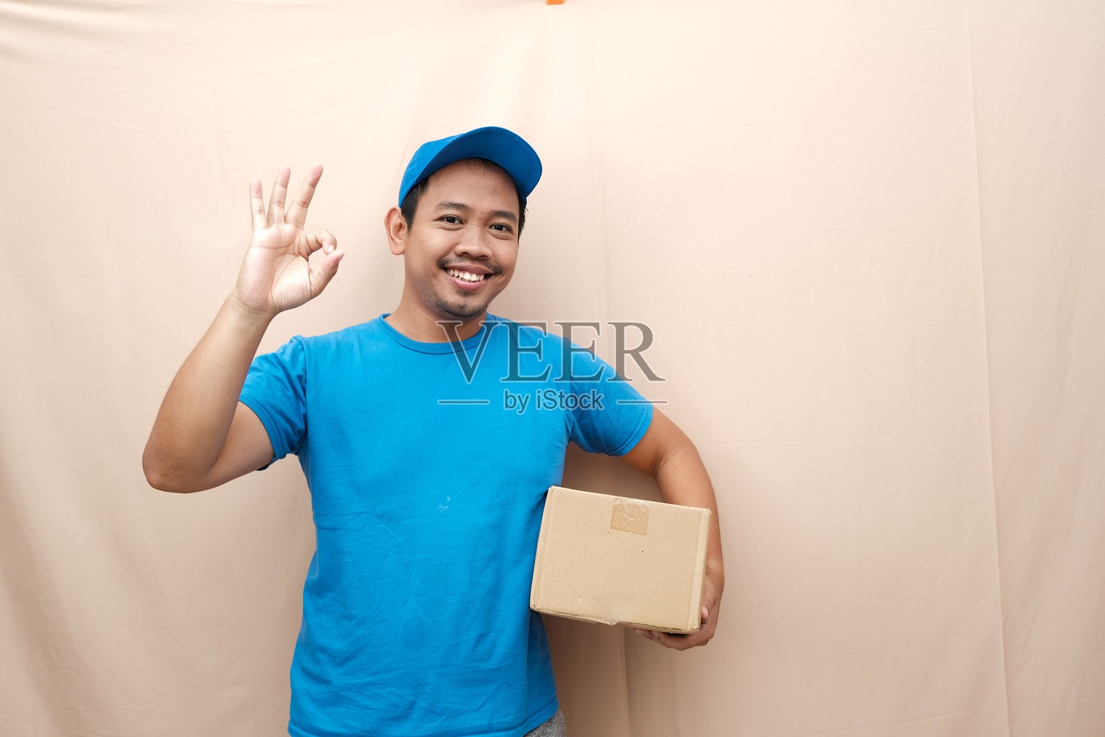 快乐的亚洲快递员穿着蓝色制服,手持包裹并做出“OK”手势表示认可。照片摄影图片