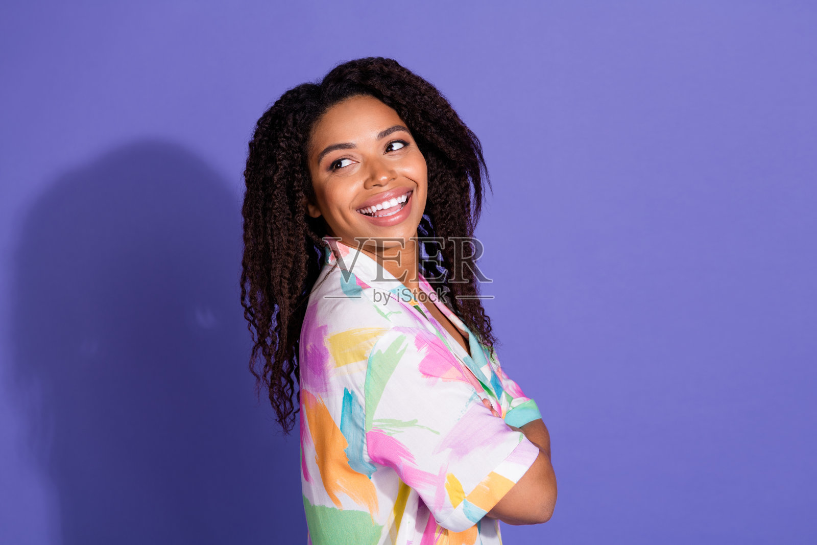 迷人的年轻女性,卷曲的黑发,穿着色彩斑斓的衬衫,在鲜艳的紫色背景前微笑。照片摄影图片