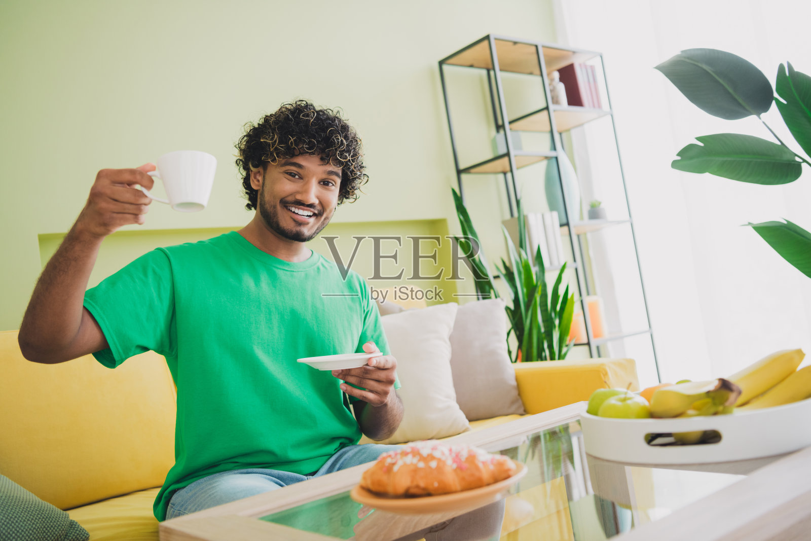 年轻男子坐在沙发上喝咖啡,穿着T恤,现代绿色室内公寓环境。照片摄影图片