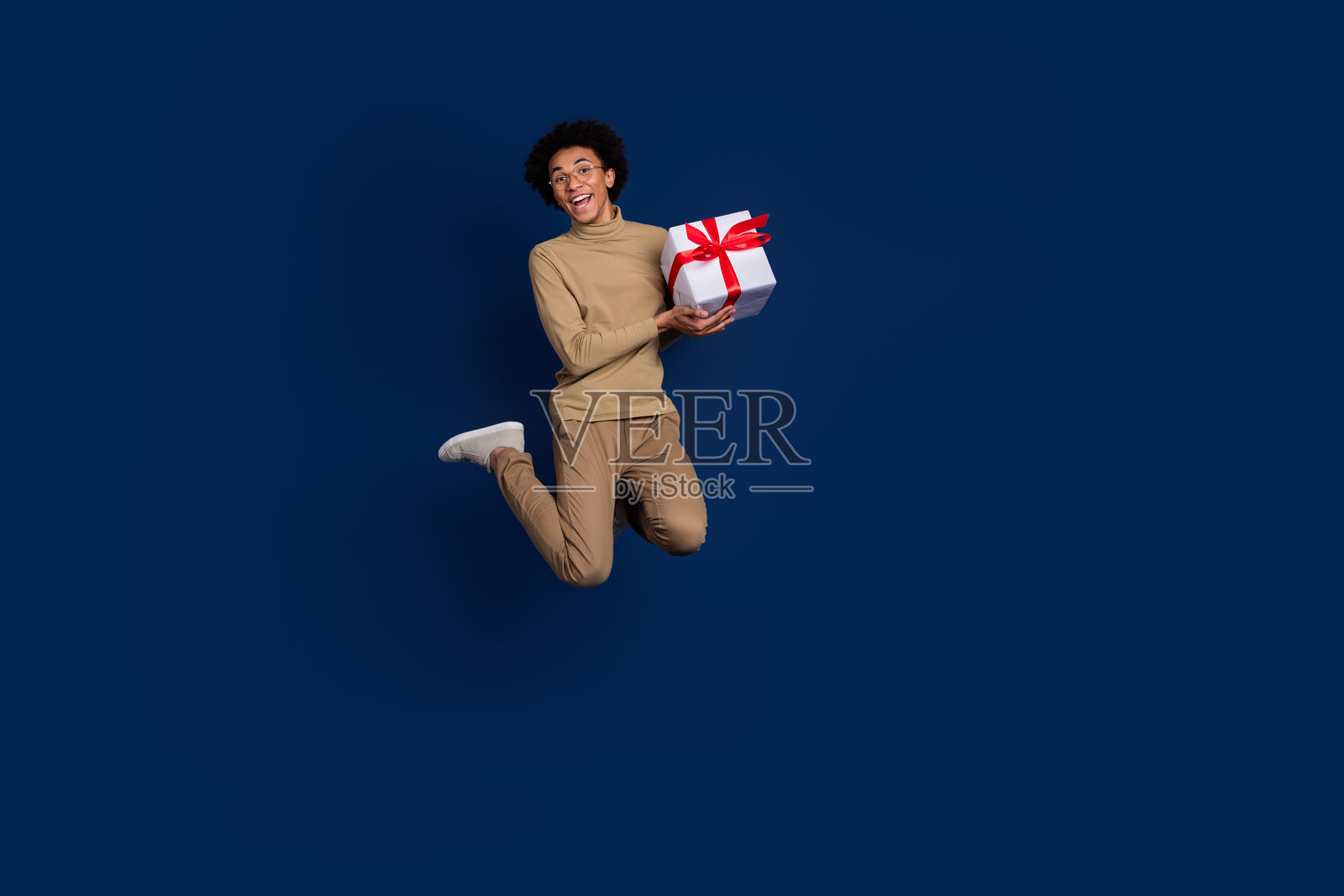 全身照,吸引人的年轻男子跳起,手持礼物,穿着时尚的米色服装,背景为深蓝色。照片摄影图片