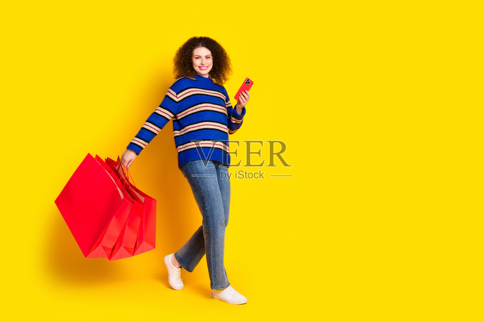 穿着针织毛衣的女孩全身照,手持购物袋和智能手机,站在黄色背景的空旷空间中。照片摄影图片