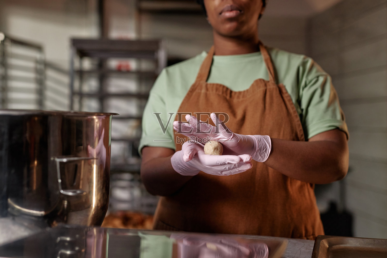 戴着丁腈手套的黑人女性面包师在商业厨房中将面团滚成球,制作面包。照片摄影图片