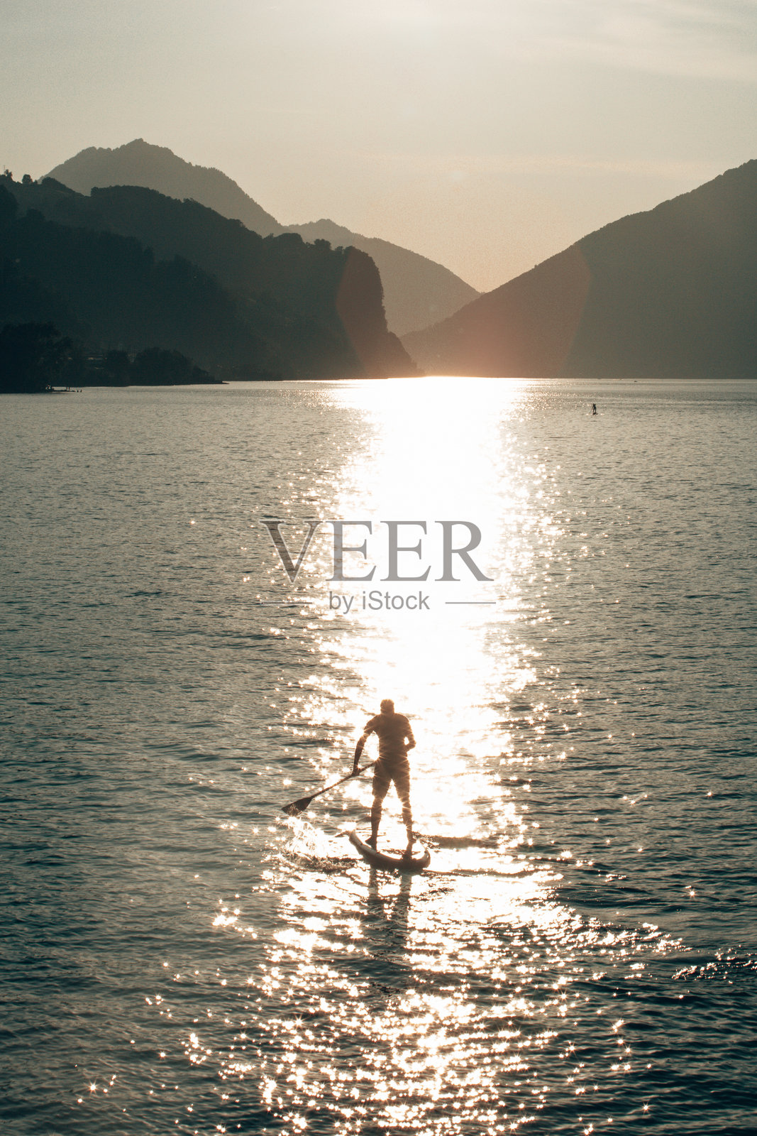 在欧洲的日落时分,山脉背景下,划水板运动者在宁静的水面上滑行。照片摄影图片