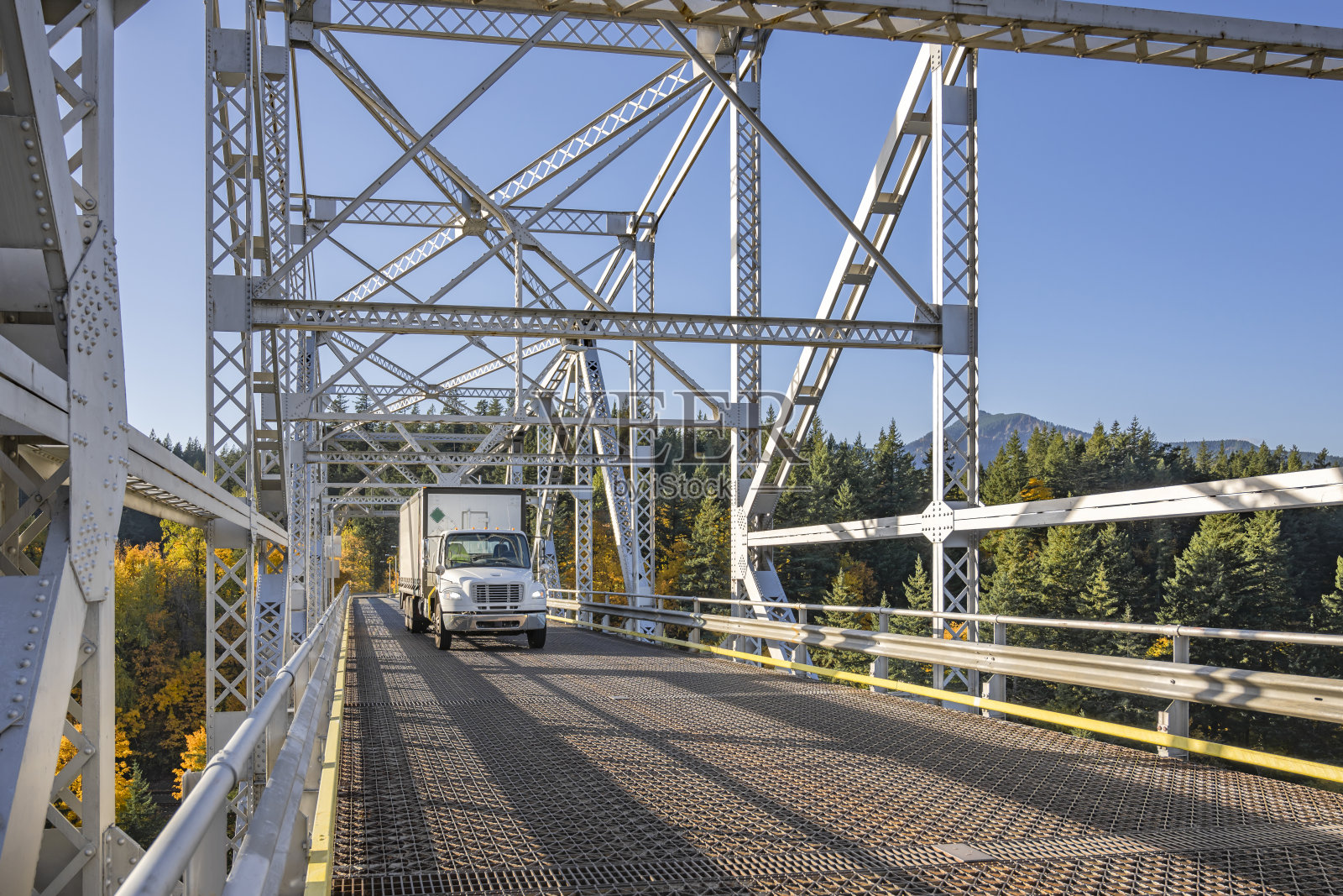 紧凑型中型白色日间驾驶室半挂卡车在哥伦比亚河上的桁架桥上运输长箱拖车货物。照片摄影图片