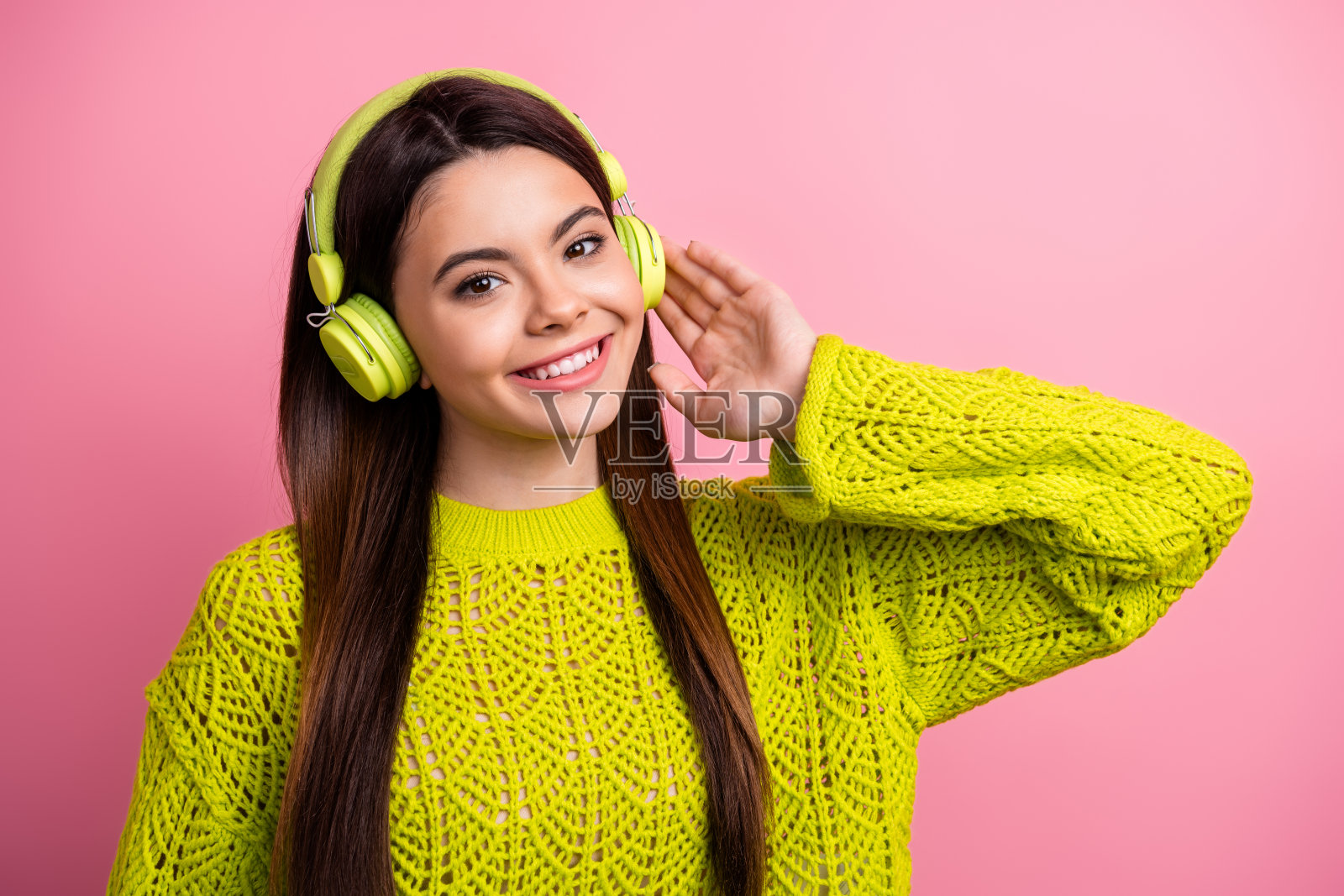 穿着黄色针织毛衣、戴着耳机的年轻女性,微笑着在鲜艳的粉色背景前摆姿势,展现出乐趣和享受。照片摄影图片