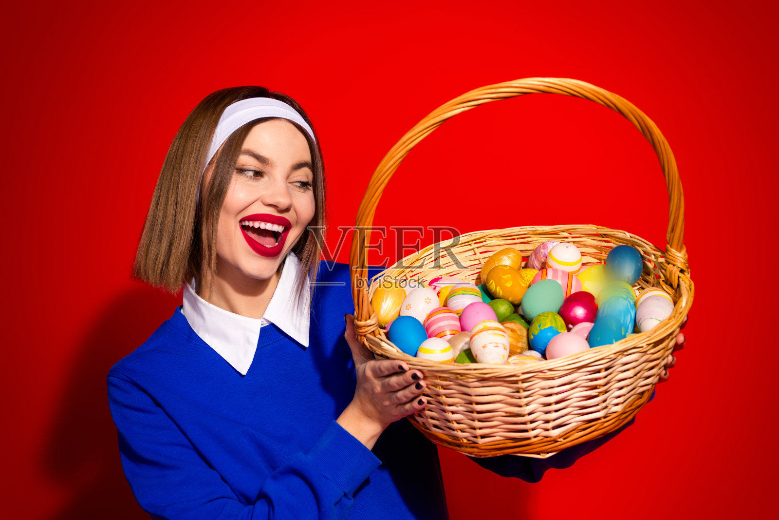 微笑的女性手持一篮五彩缤纷的复活节彩蛋,背景为鲜艳的红色。照片摄影图片