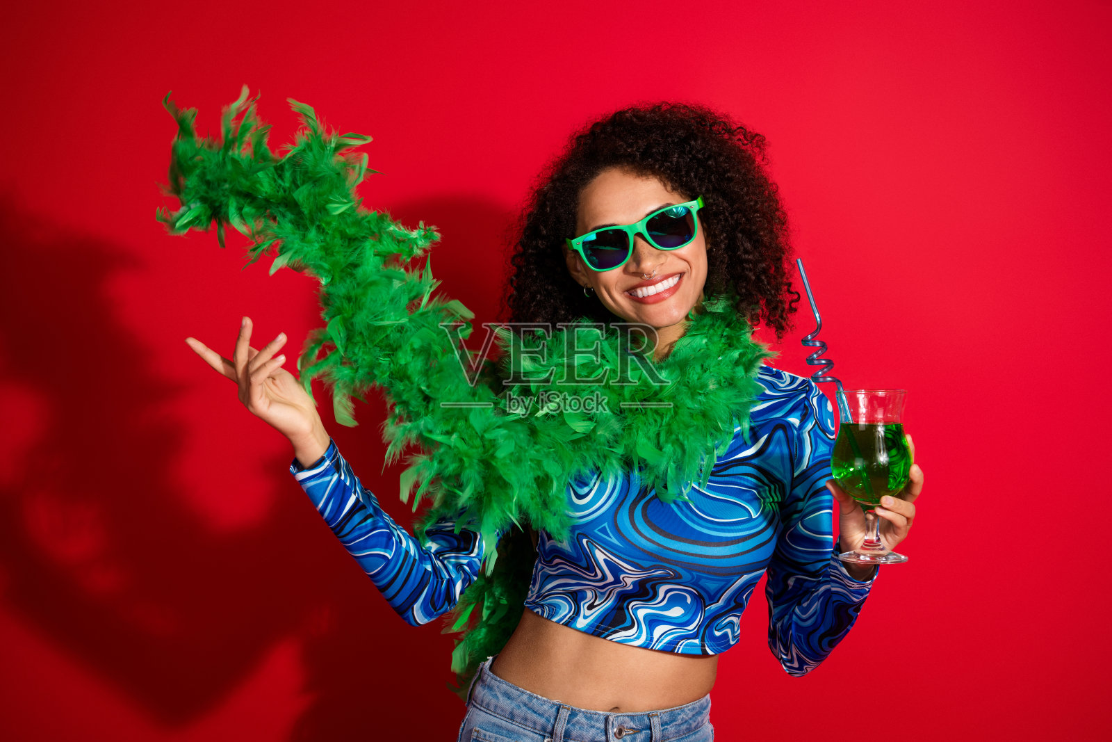 年轻女性穿着时尚的绿色羽毛围巾和太阳镜,手持饮料,面带微笑,背景为鲜艳的红色,展现出乐趣和自信。照片摄影图片