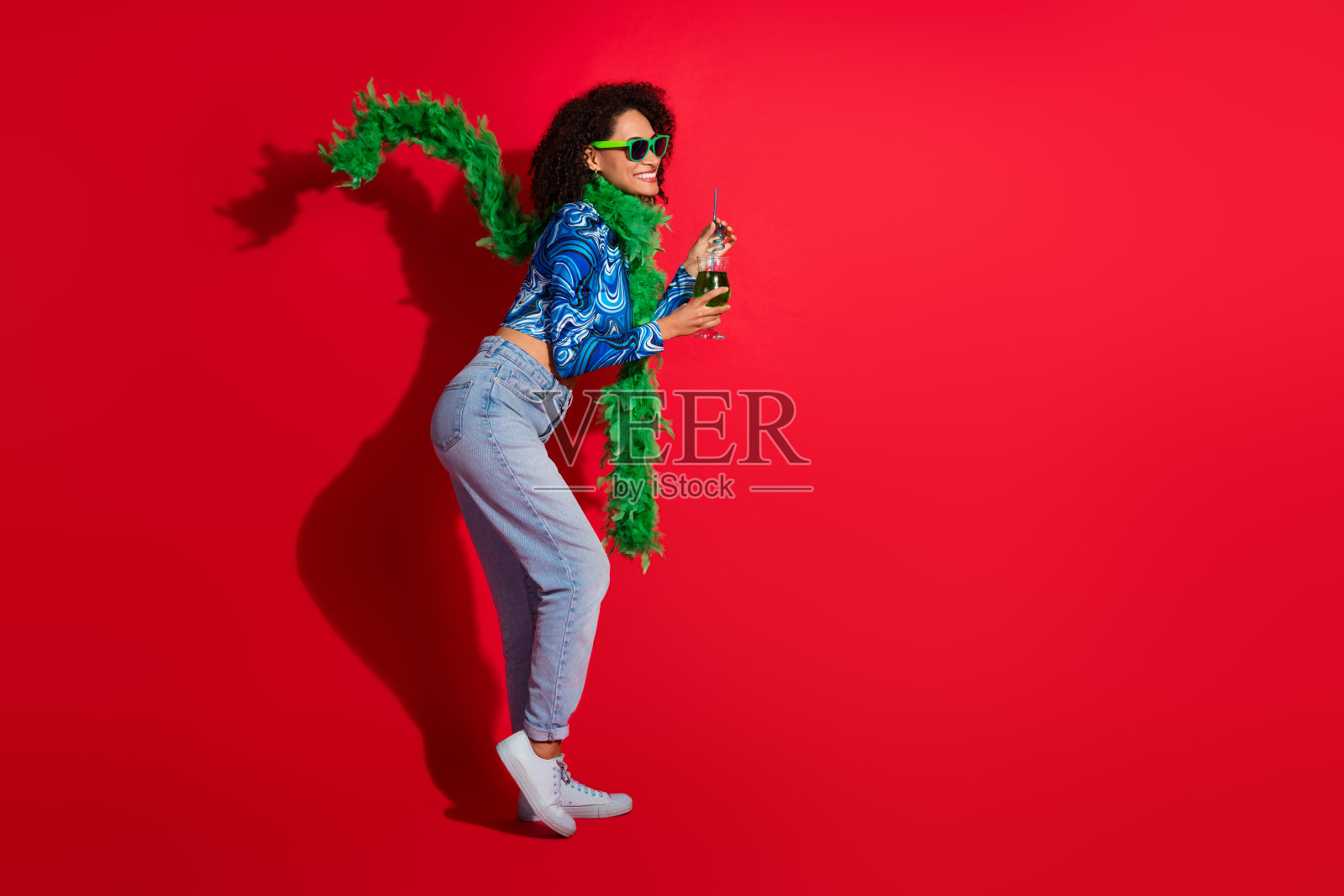 时尚女性在红色背景前摆姿势,穿着色彩斑斓的服装和太阳镜,手中拿着饮料,搭配绿色羽毛围巾。照片摄影图片